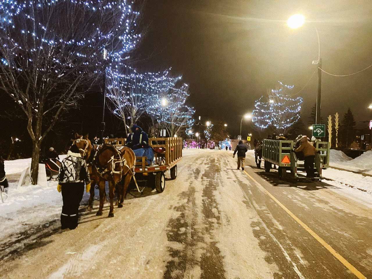 Street at night with snow-covered ground, horse-drawn carriages, people, and trees adorned with blue holiday lights.