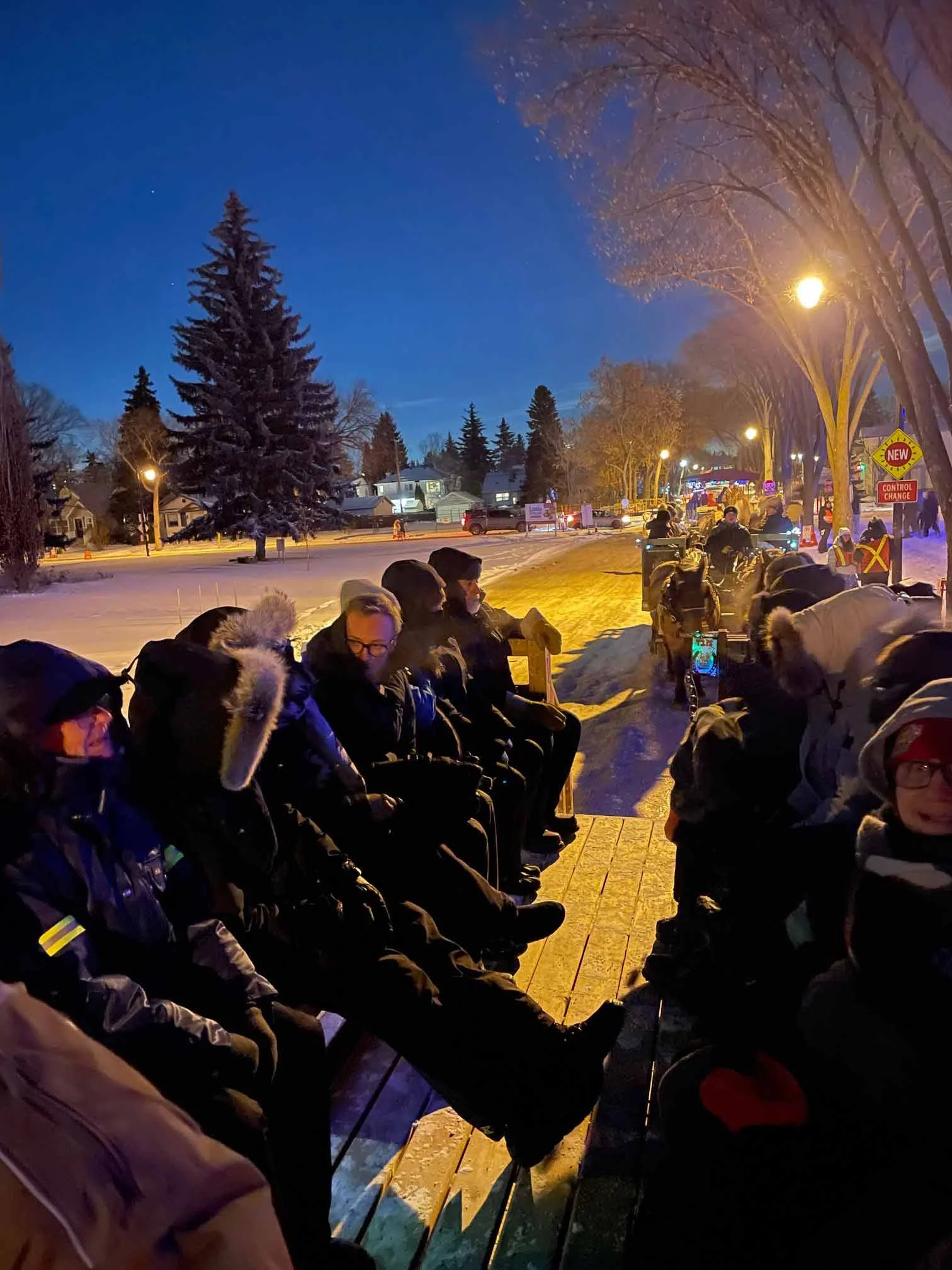 Group of people sitting in chairs outdoors on a snowy evening, illuminated by streetlights, with snow-covered trees and houses in the background.