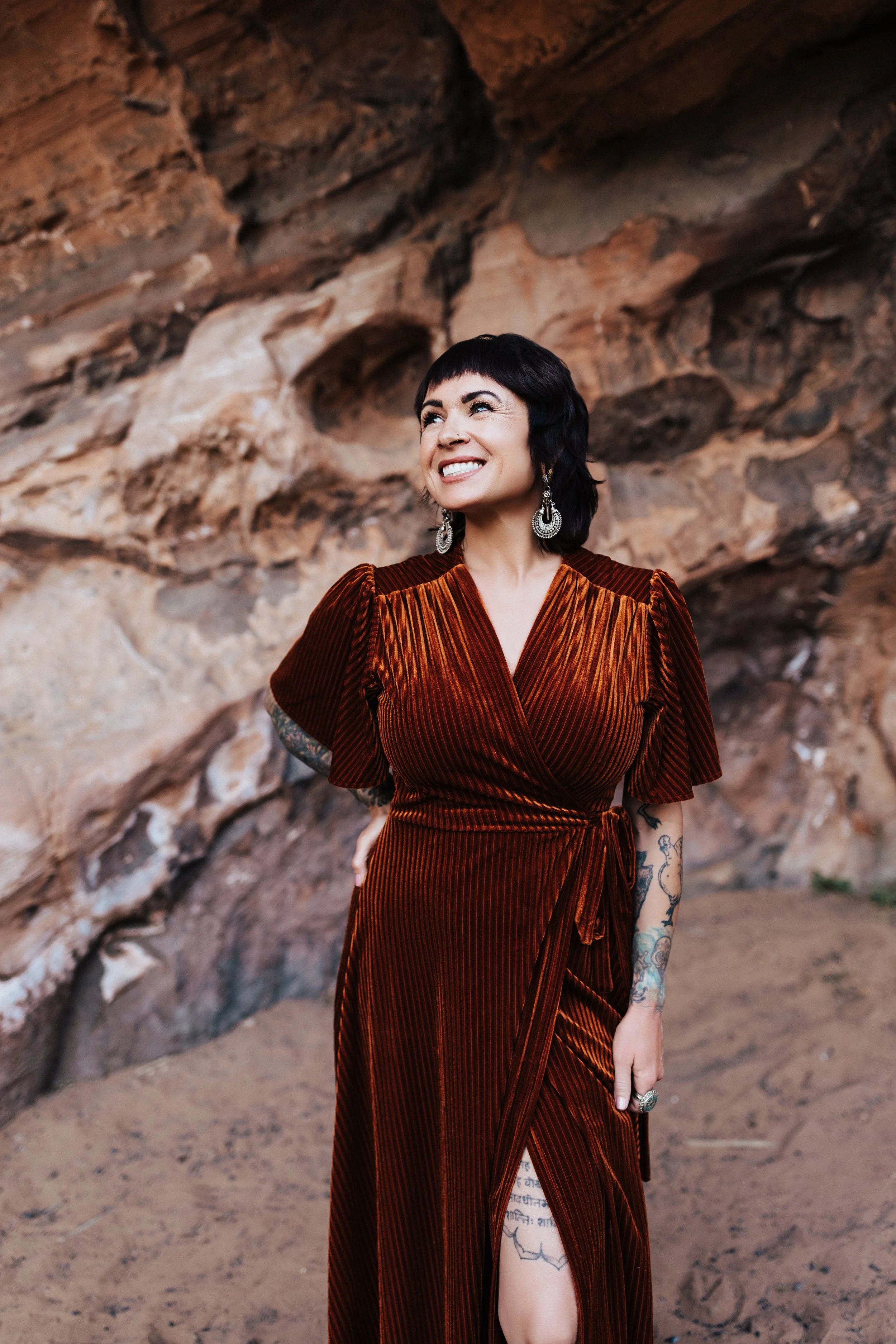 A woman with short black hair and tattoos, wearing a rust-colored velvet dress with puffed sleeves, standing outdoors in front of a rocky background, smiling and looking to her left.