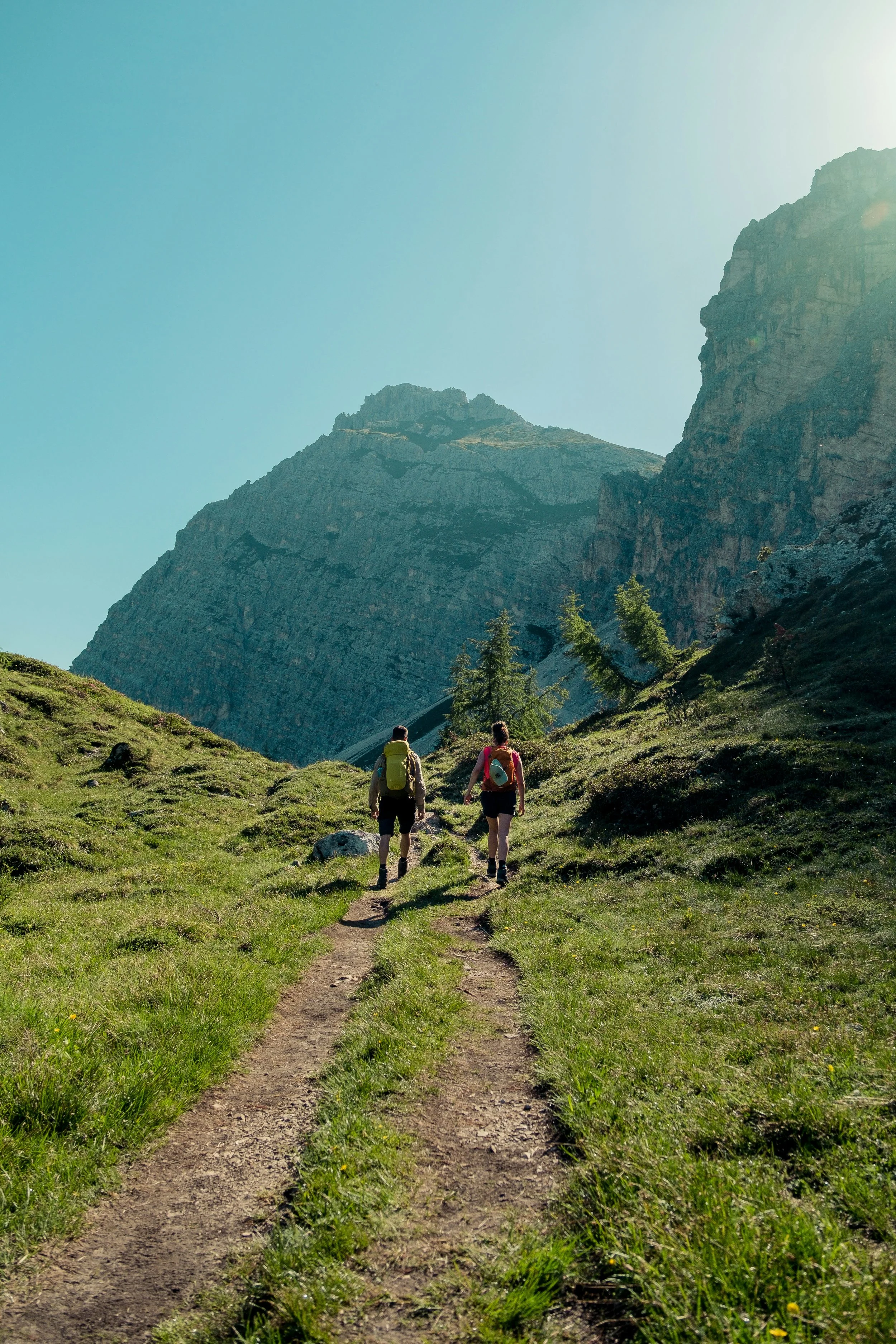 Two hikers with backpacks walking on a dirt trail through a lush green mountainous landscape.