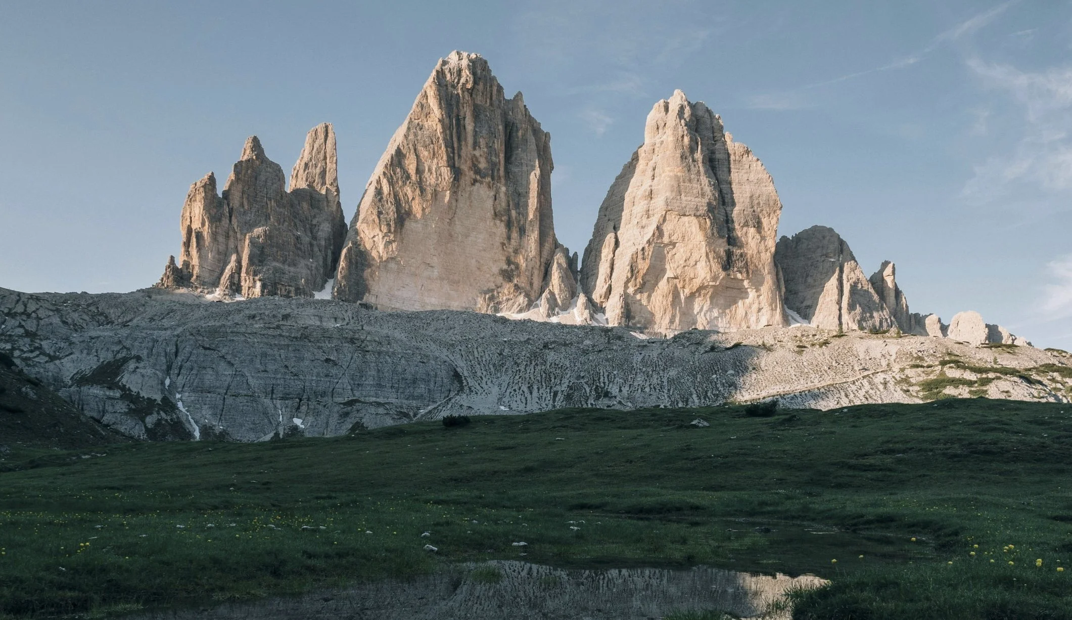 A mountain range with three tall, jagged peaks rising above a grassy foreground and a small pond reflecting the sky.