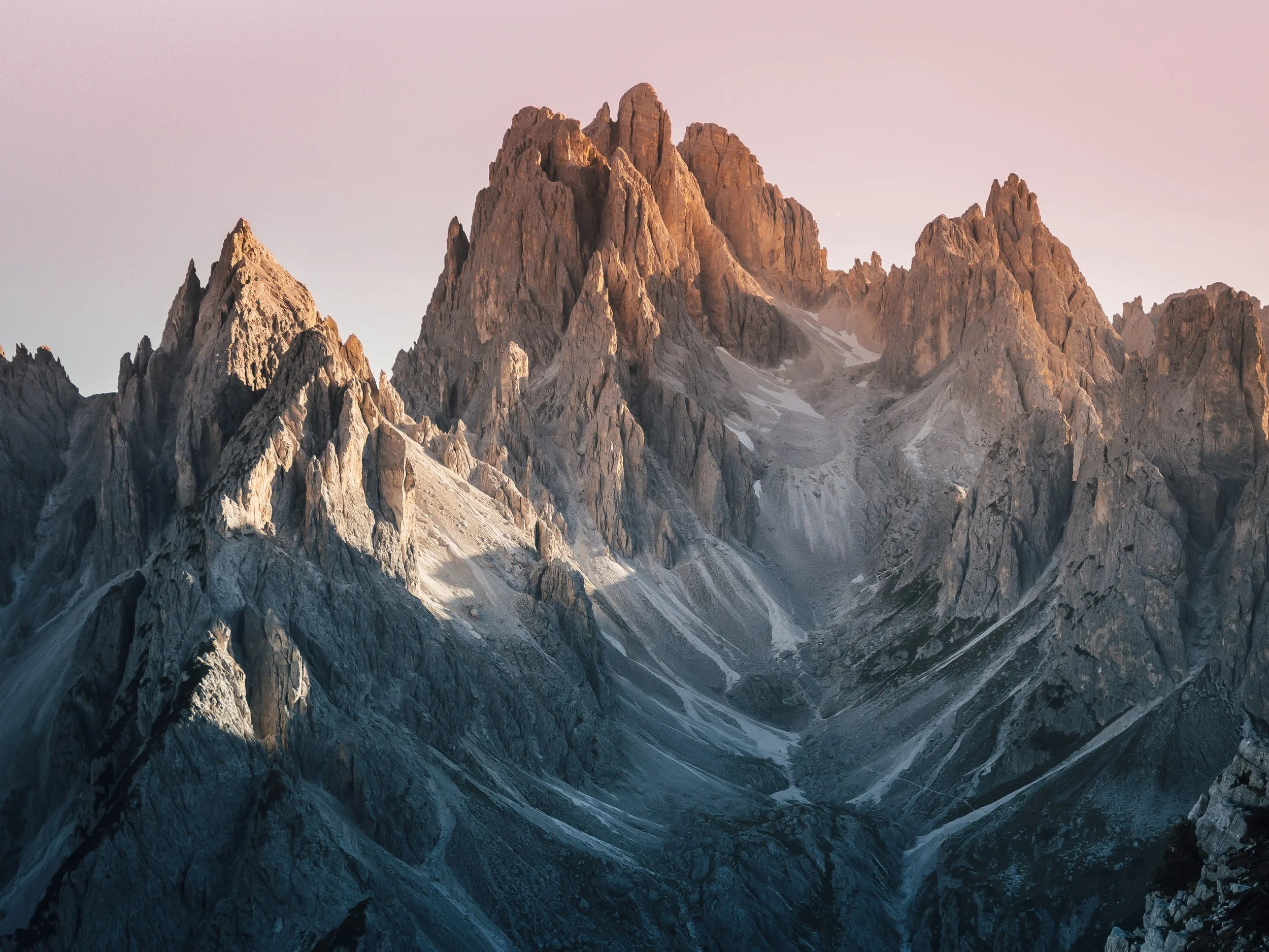 Sunlight illuminating rugged mountain peaks with snow patches on a pinkish sky background