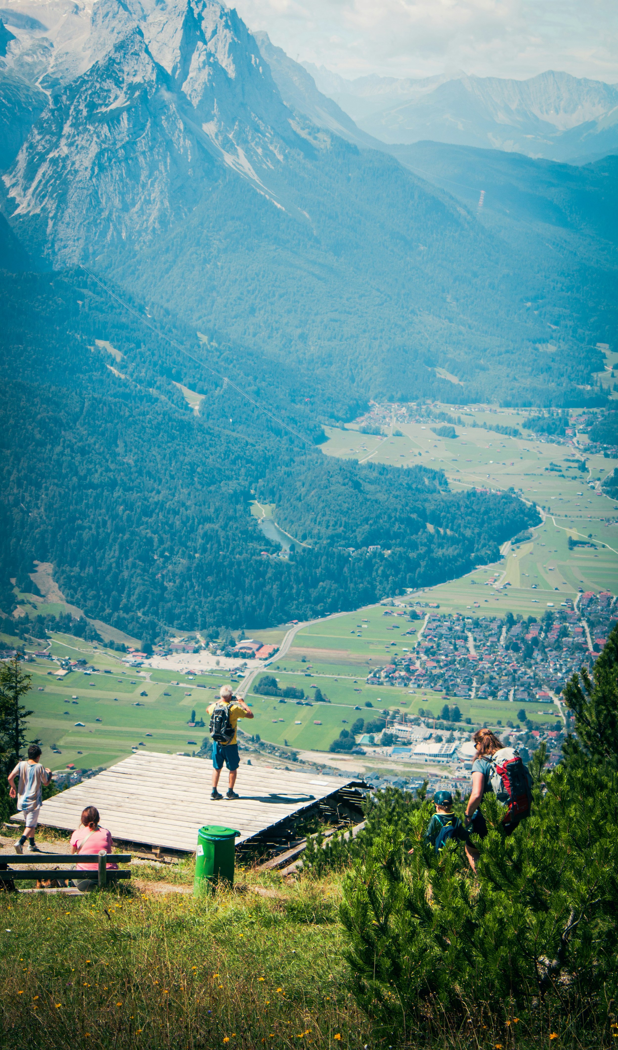 Group of hikers on a mountain viewpoint overlooking a valley with green fields, a river, and a small town, surrounded by forested mountains.