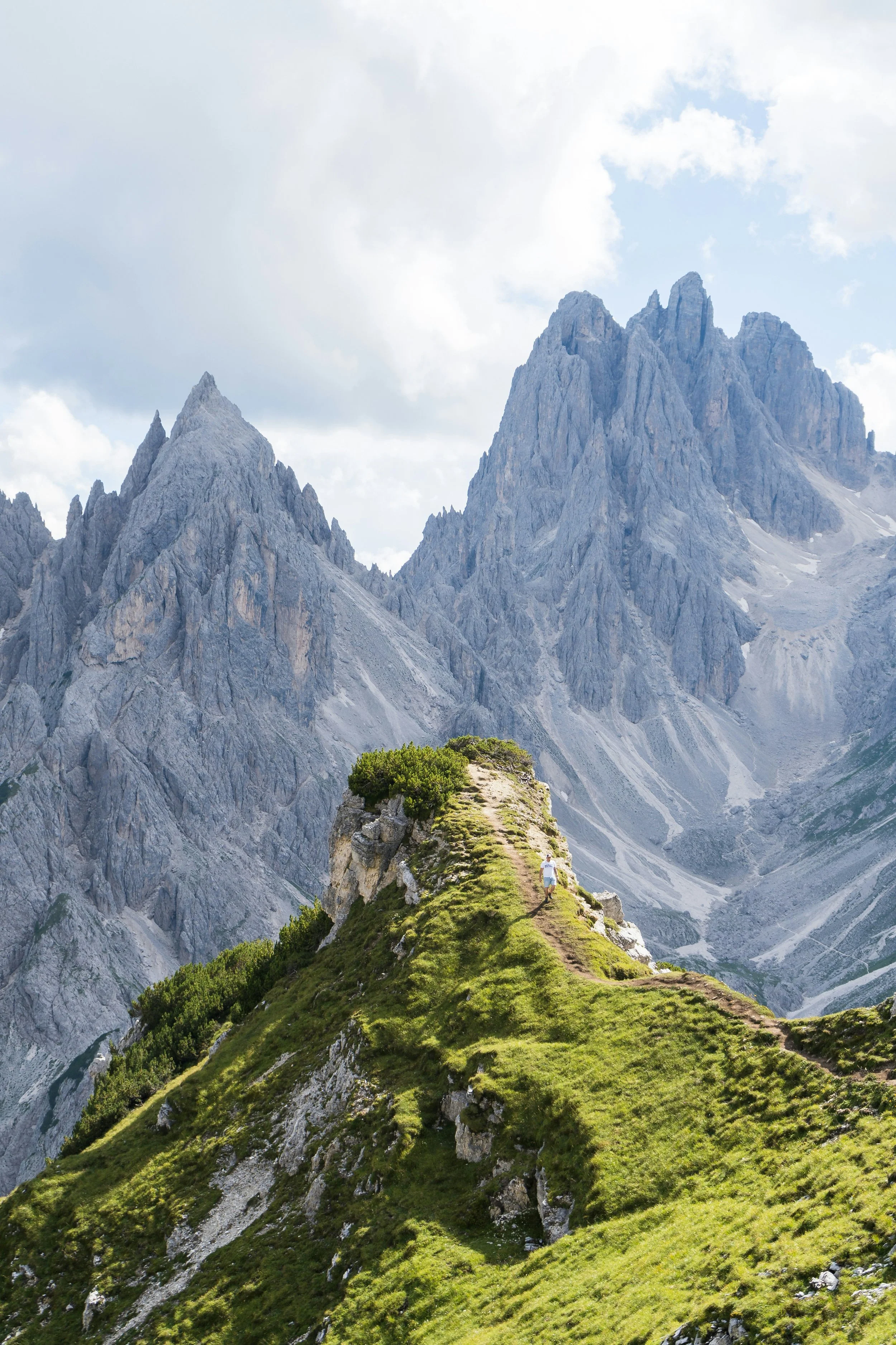 A person walking on a grassy trail along a small ridge with steep rocky mountain peaks in the background under a cloudy sky.