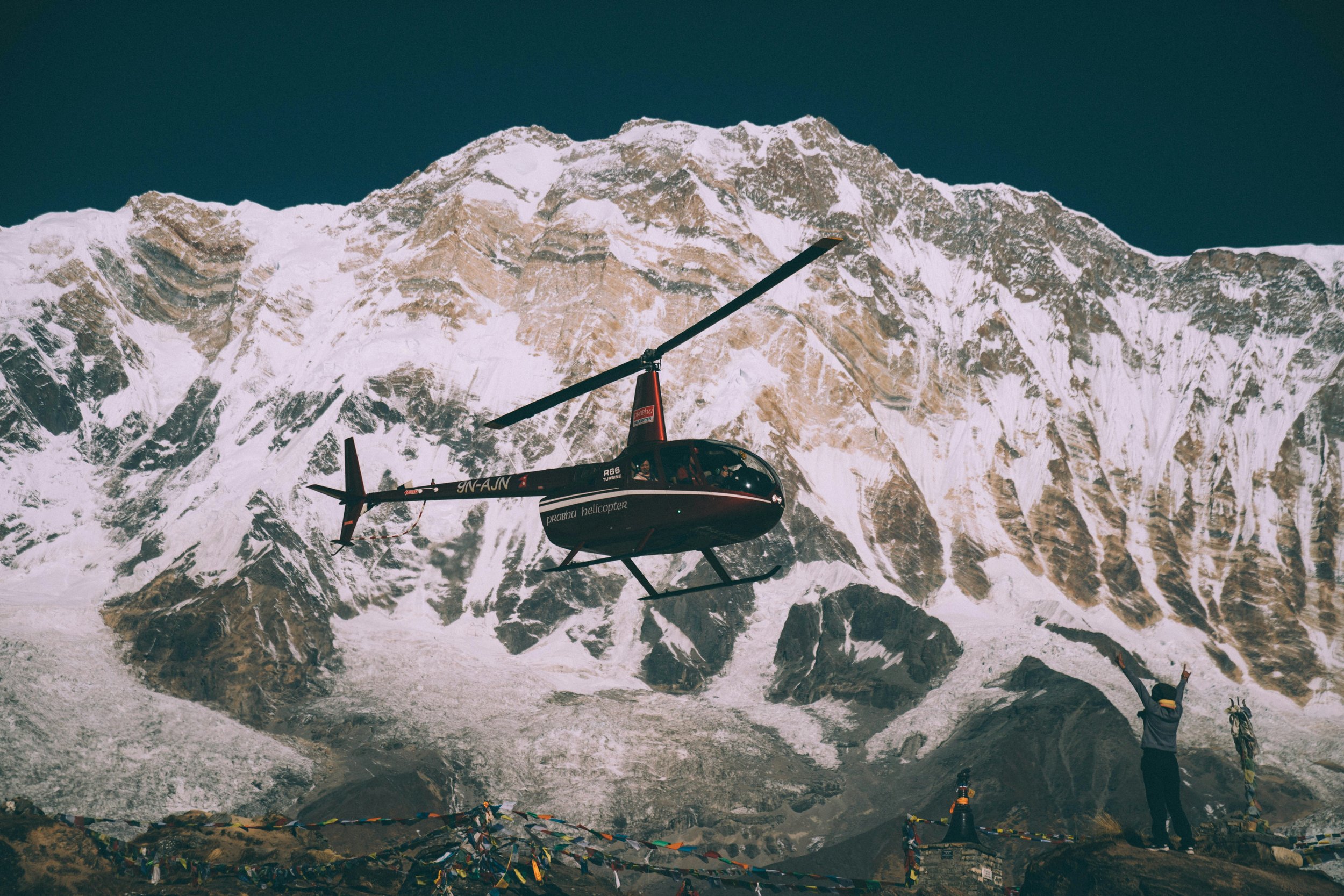 A helicopter flying near snow-covered mountain peaks with a person standing on the ground below, raising their arms in celebration.