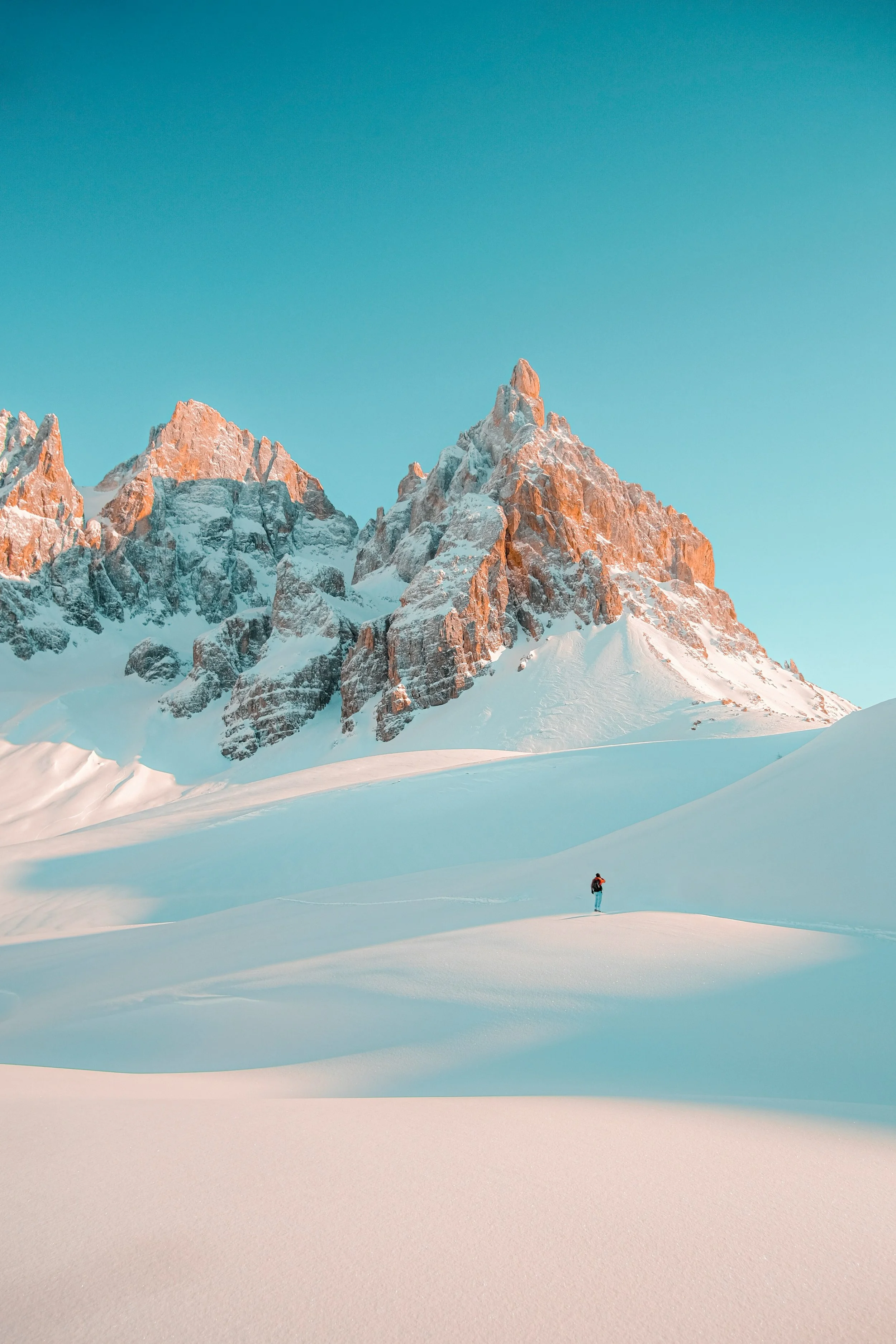 A lone hiker walks across snow-covered terrain in front of towering snow-capped mountains under a clear blue sky.