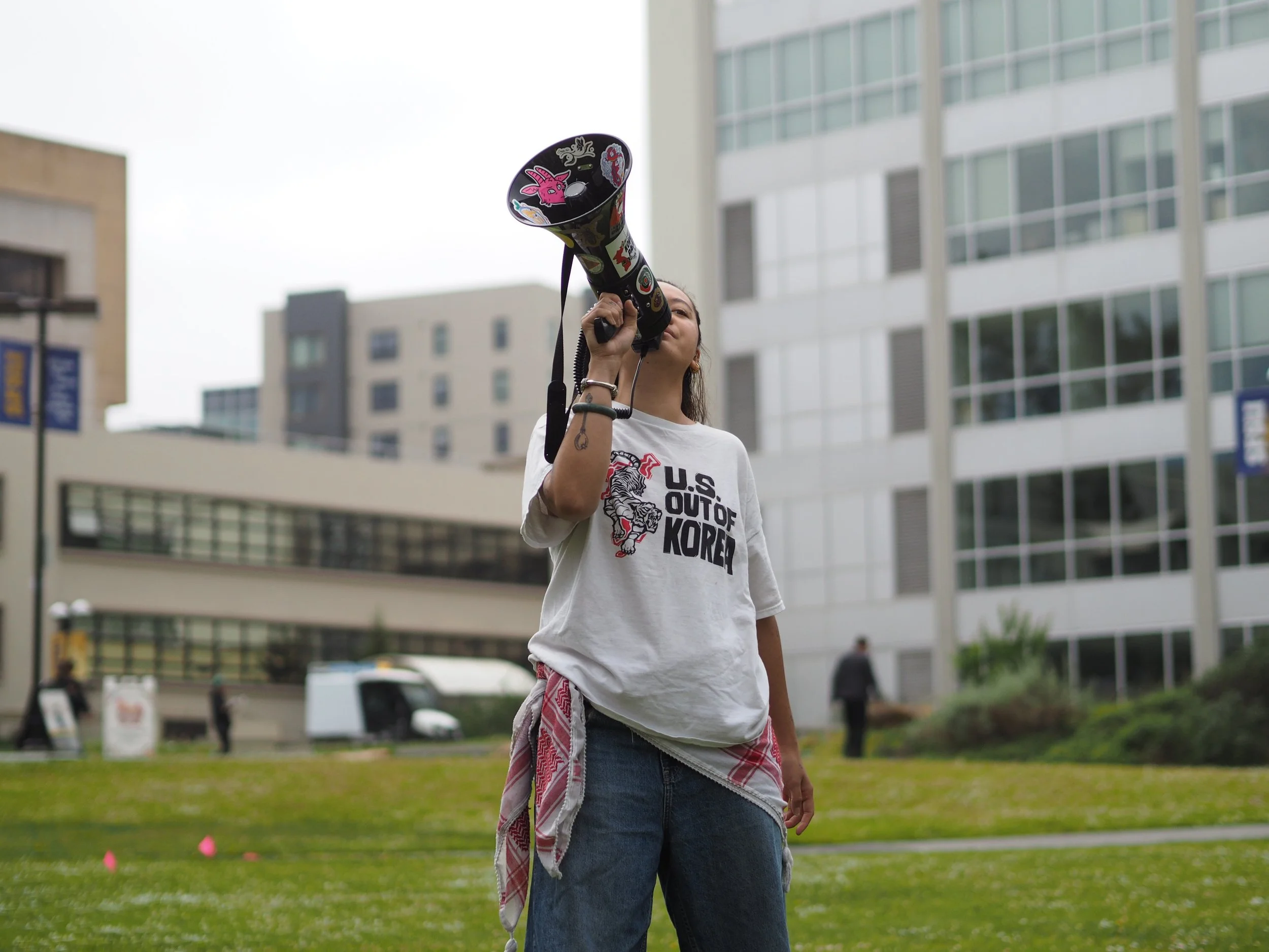 An Asian American person stands in the foreground with a megaphone at their mouth, raised to the sky. They wear a T-shirt that reads "U.S. out of Korea" and a keffiyeh around their waist. 