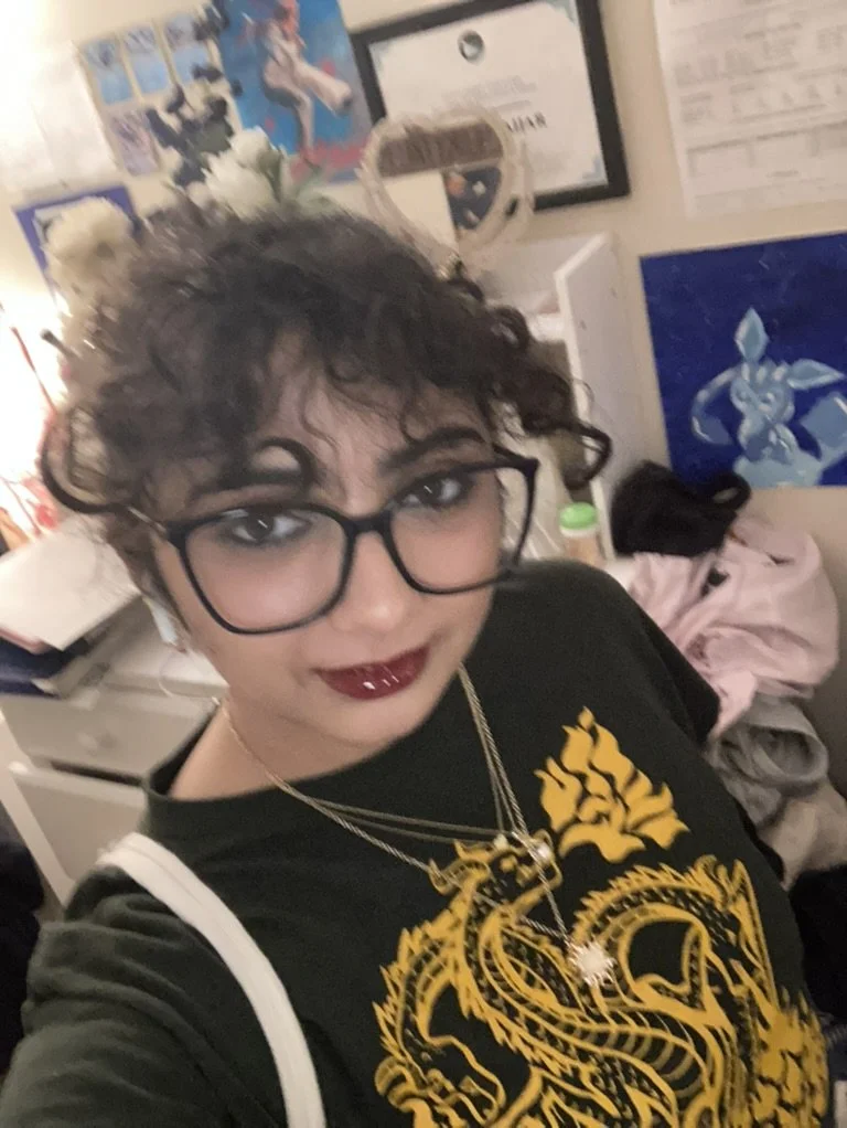 An Arab person with curly brown hair and glasses, wearing a black T-shirt.