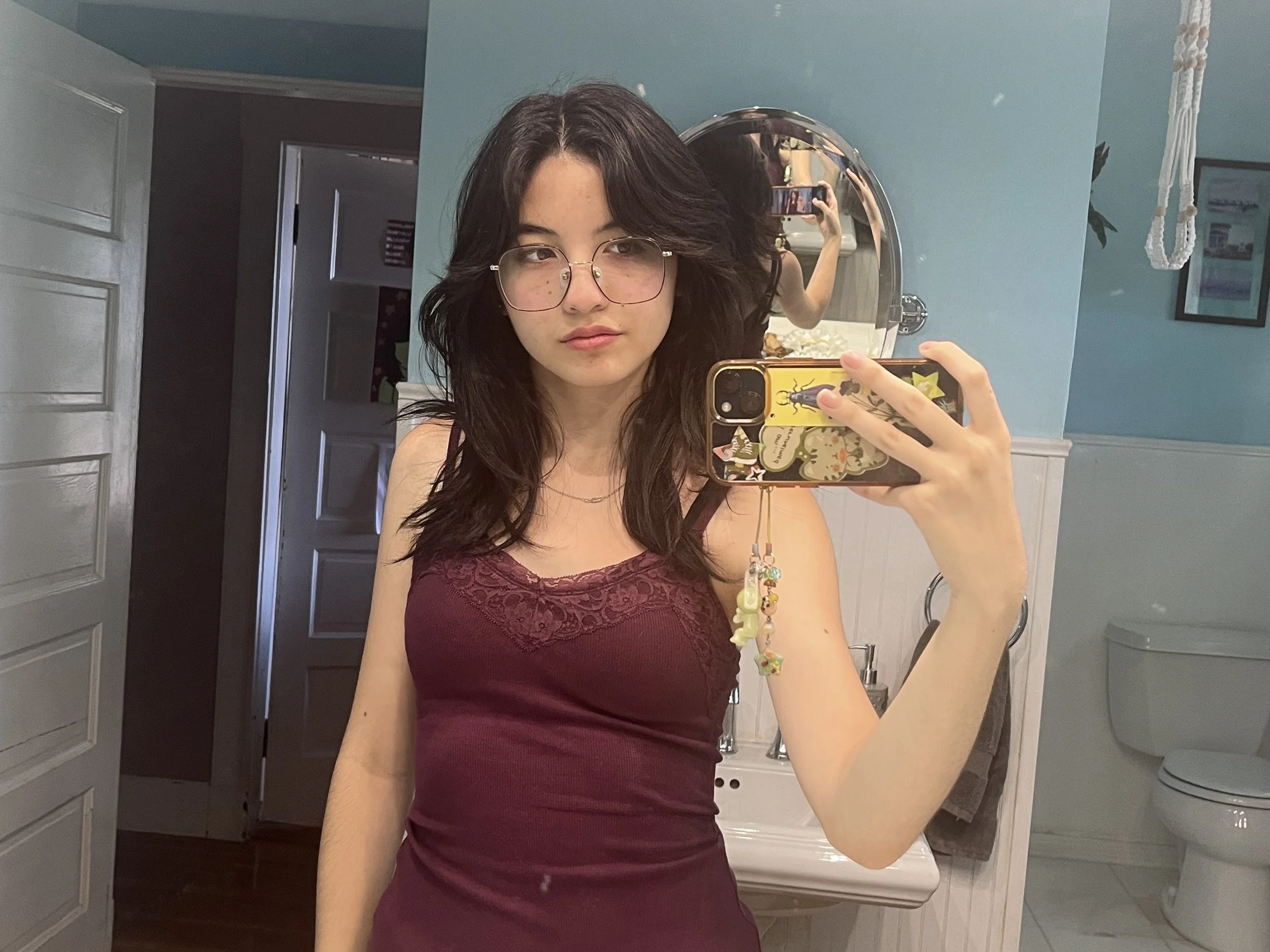 An Asian American person with medium-lng brown hair and glasses, wearing a maroon tank top takes a mirror selfie.
