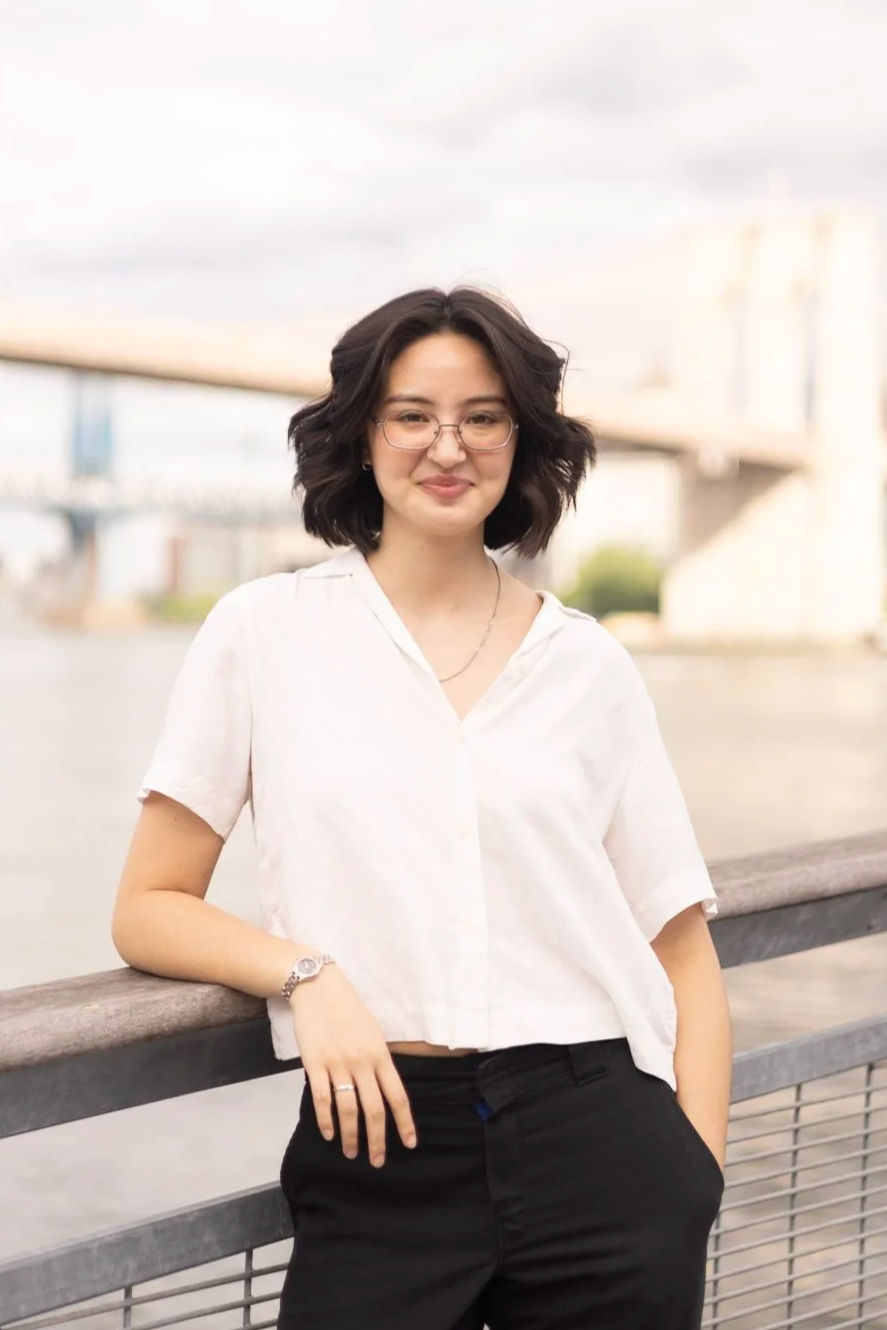 An Asian American person leans on a railing. They have a white shirt and black pants and shoulder-length brown hair.