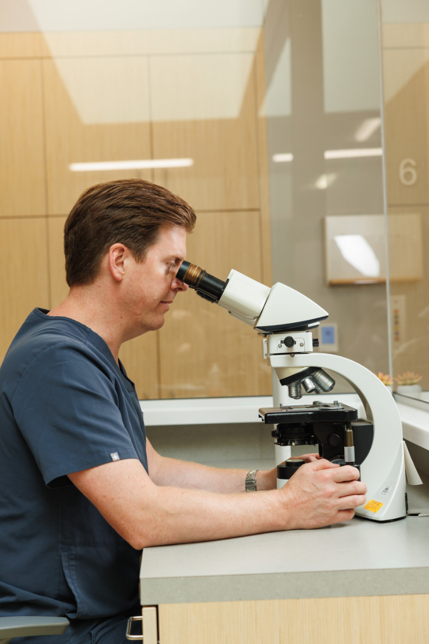 Dr. Chad Prather in medical scrubs using a microscope in a laboratory with wood-paneled walls.