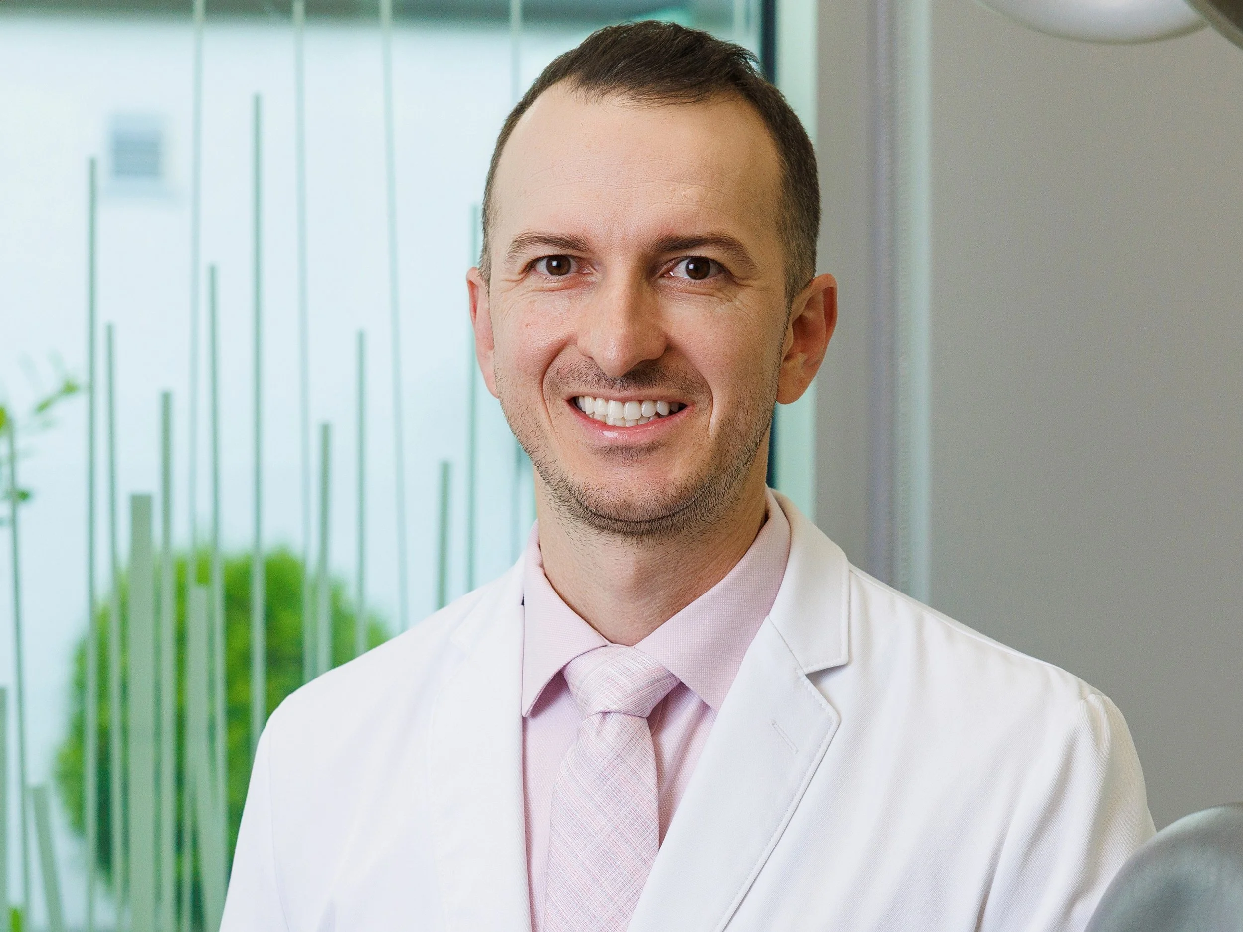 Dr. Adam Carter with short brown hair and light skin is smiling at the camera. He is wearing a white shirt and a light pink tie. The background shows a modern office or clinic with green plants and glass walls.