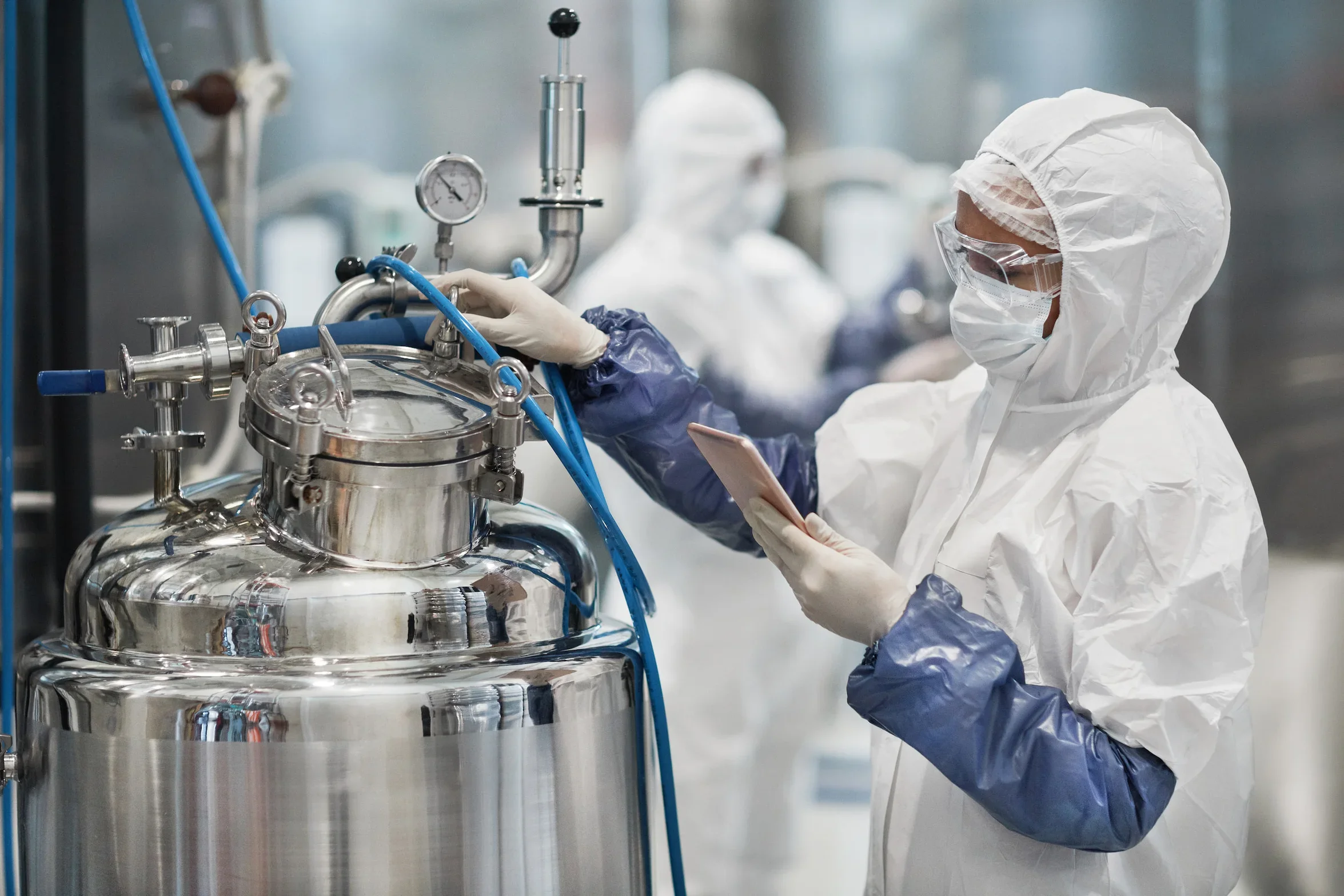 A technician in full PPE inspects a stainless steel process vessel in a pharmaceutical cleanroom, illustrating solvent handling in drug manufacturing