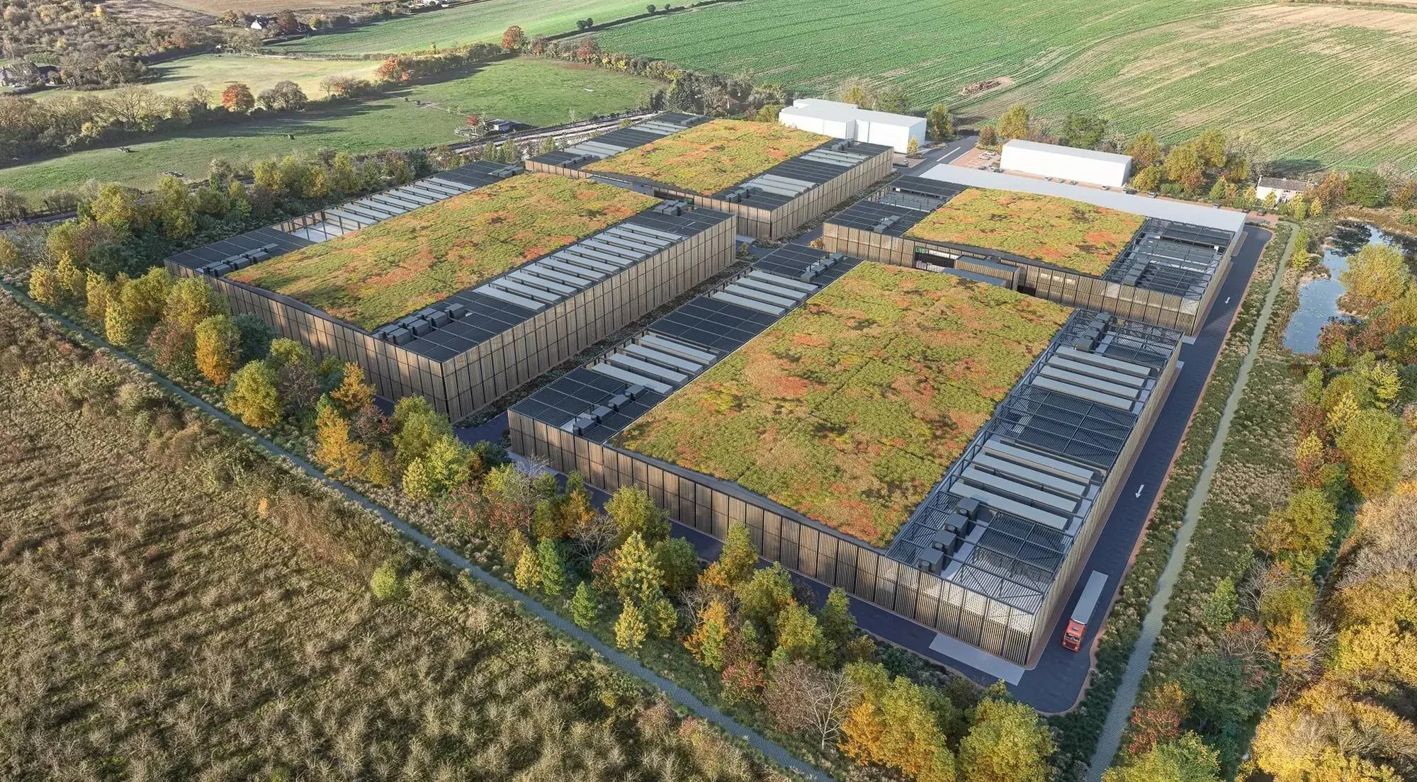 Aerial view of a data centre campus by Bryden Wood, with green roofs, rooftop plant, timber-toned facades and tree planting that screens the buildings from surrounding fields, woodland and a pond.