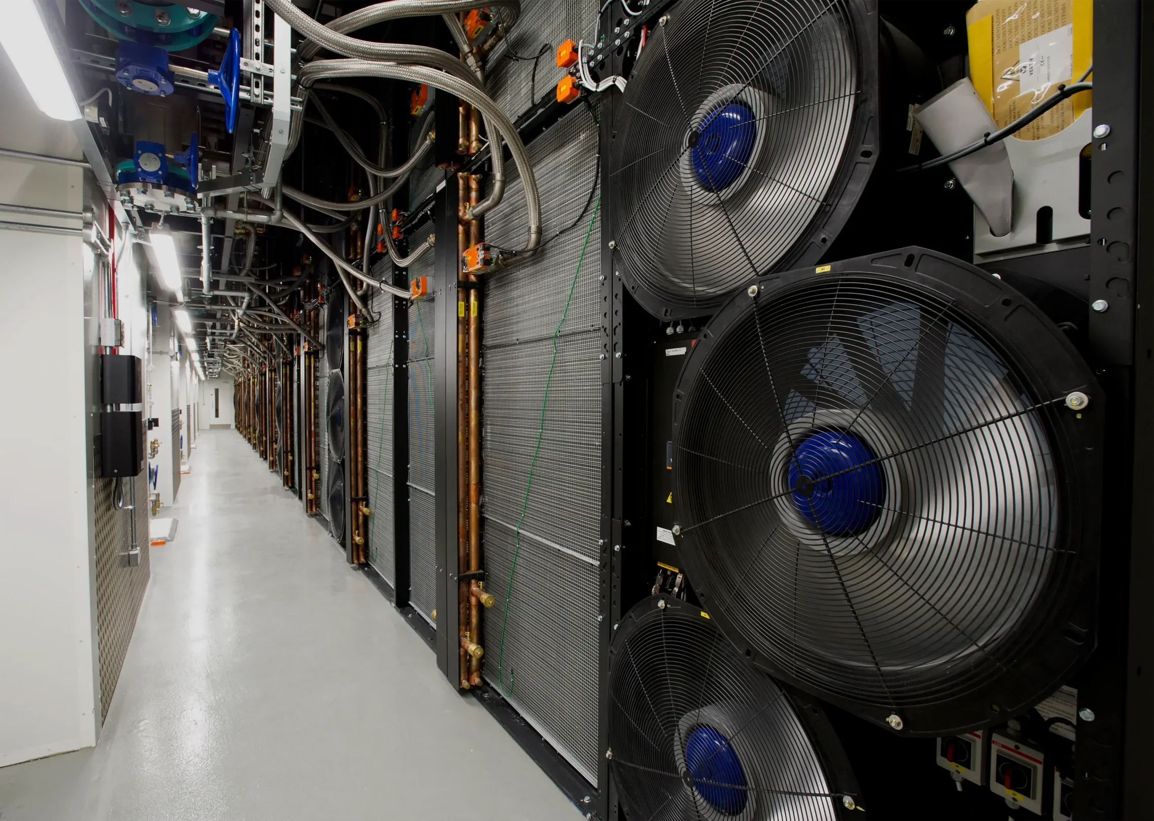 Internal corridor of a data centre showing a row of large fan wall cooling units with copper pipework and overhead MEP services, illustrating the scale of mechanical infrastructure required in high-density facilities