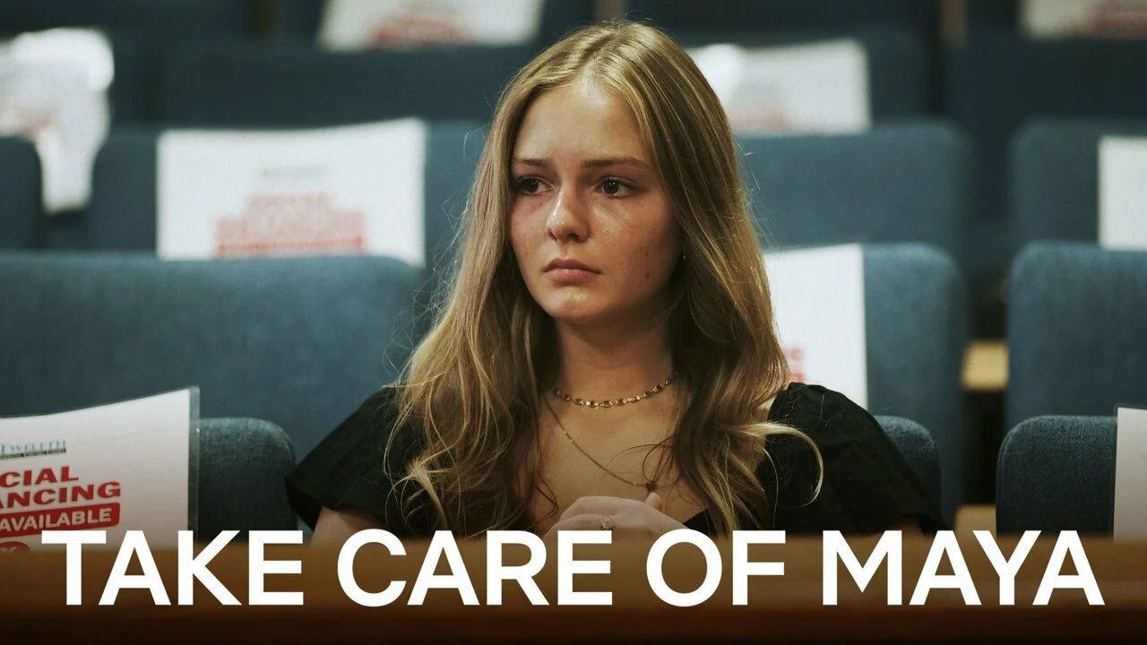 A young woman sitting in an auditorium, appearing emotional, with the text 'Take Care of Maya' overlaid at the bottom. Background shows empty seats with social distancing signs.