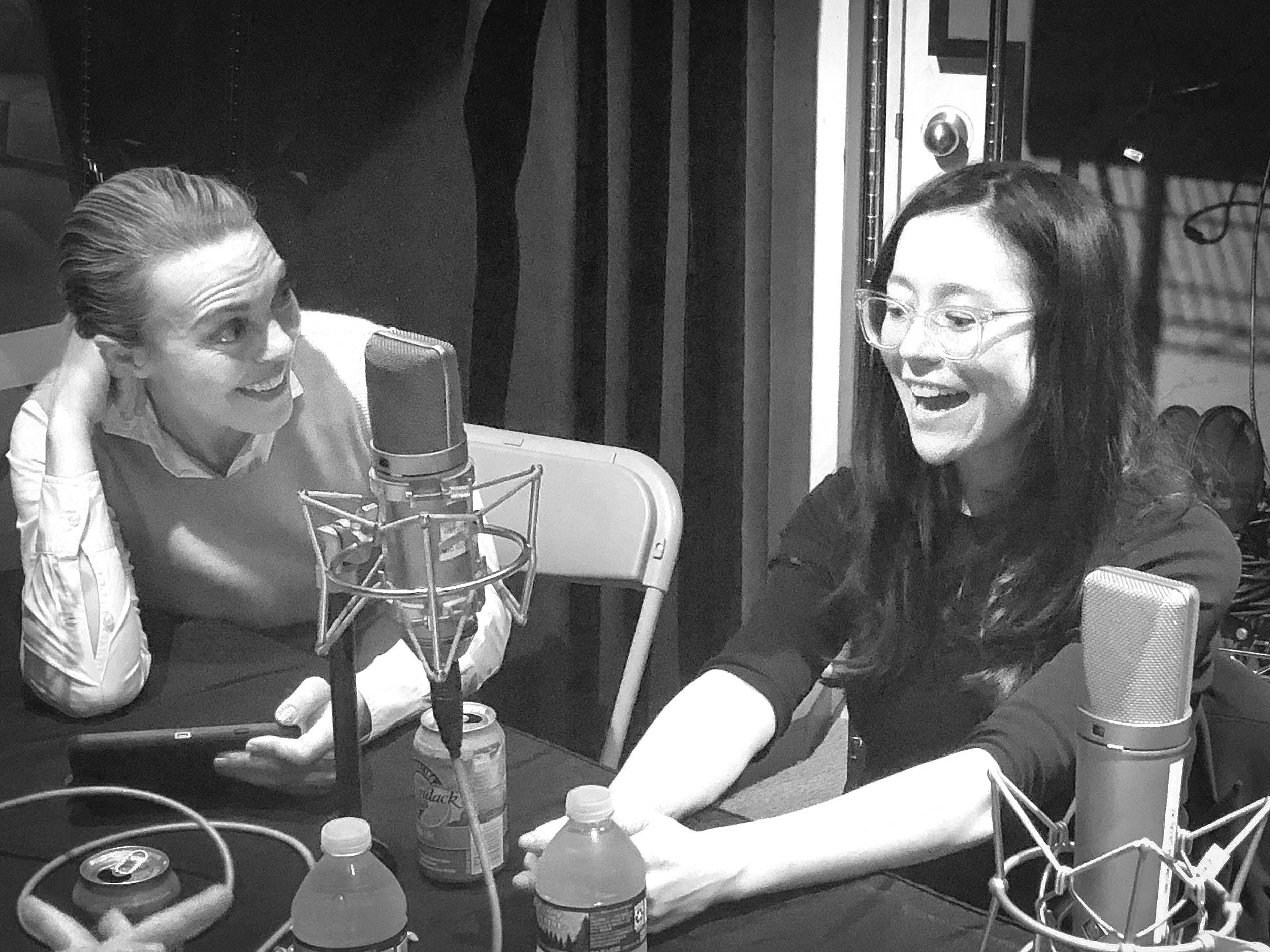 Two individuals talking and laughing in a recording studio with microphones, cans, and bottles on the table.