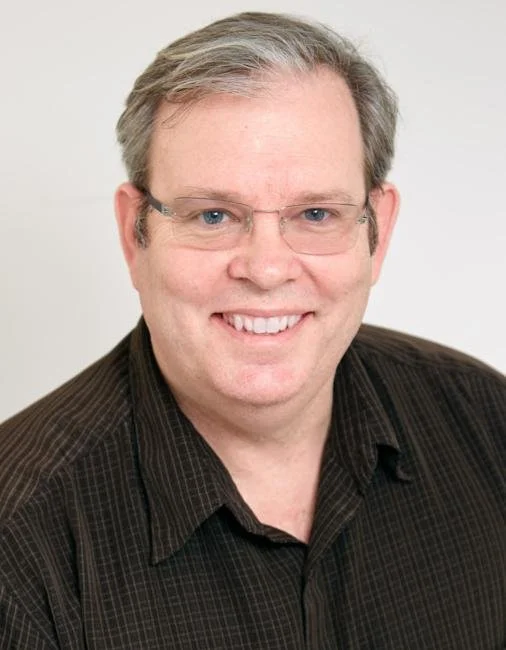 Portrait of a smiling person wearing glasses and a dark shirt, with light-colored hair.