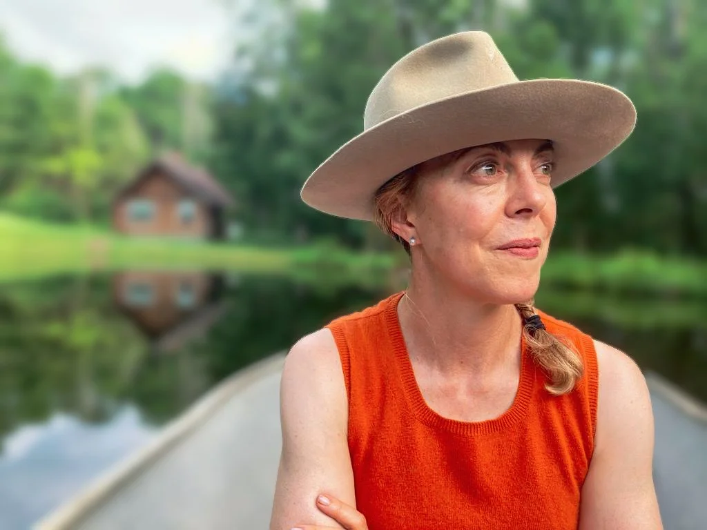 Woman in a hat and orange sleeveless top sitting outdoors with a pond and cabin in the blurred background.