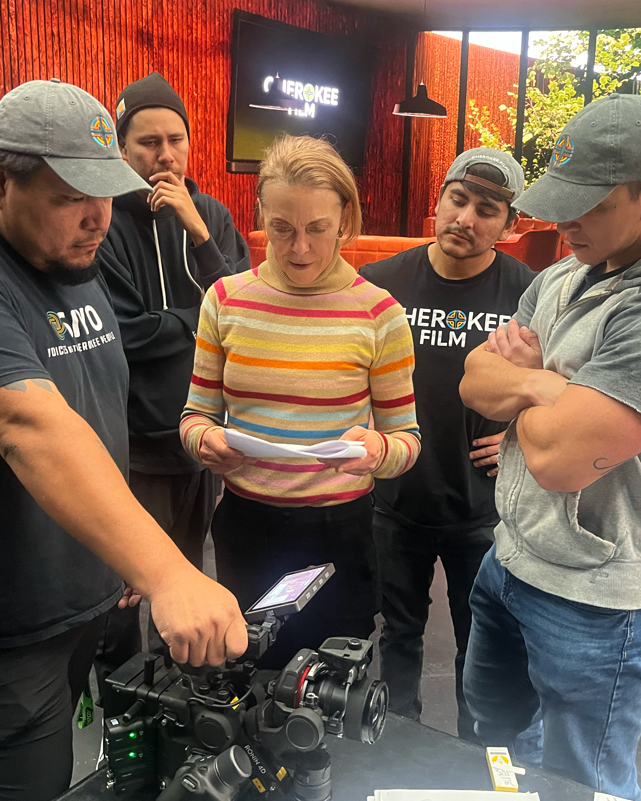 Group of people gathered around a table with camera equipment, reviewing notes in a production setting, with "Cherokee Film" visible on a monitor in the background.