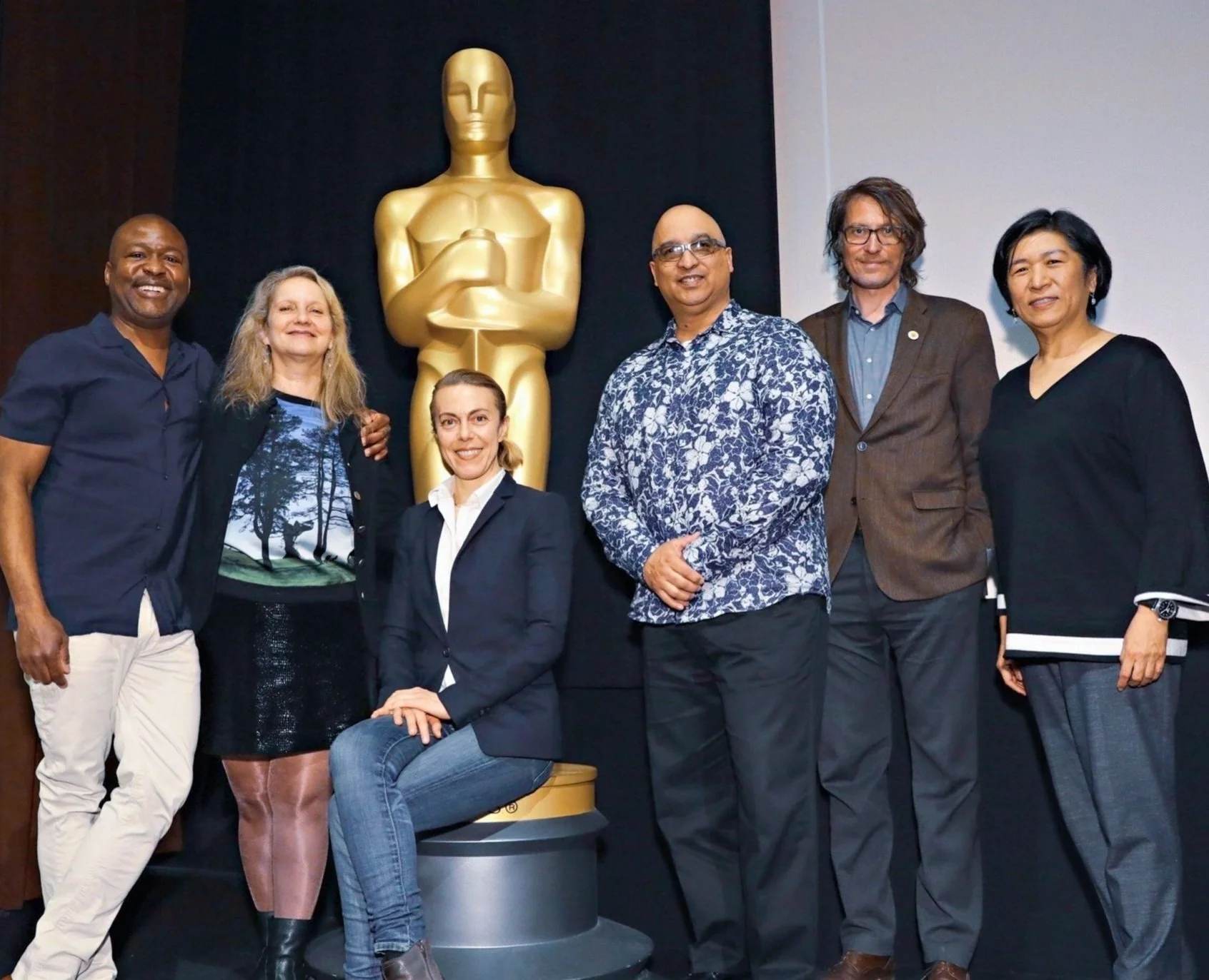 A group of six people stand in front of a large gold Oscar statue, posing for a photo. They are dressed in business casual attire, with one person seated in the center. The setting appears to be indoors, possibly at an event or ceremony.