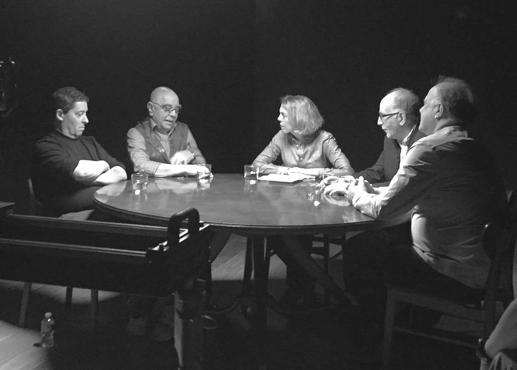 Five people seated at a round table engaged in discussion, lit by overhead lighting, glass of water on table.