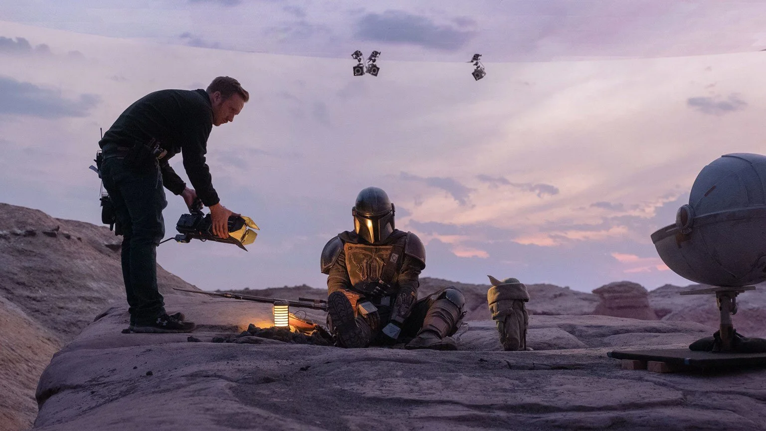 Behind-the-scenes of a film set with a person adjusting lighting on a rocky landscape at sunrise or sunset, featuring a character in armor and a fictional creature.