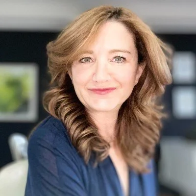 Person with long, wavy hair, smiling, and wearing a dark top, indoors.