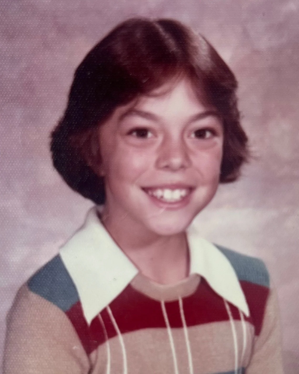 Vintage portrait of a smiling child with short hair wearing a striped sweater and a collared shirt, set against a neutral backdrop.