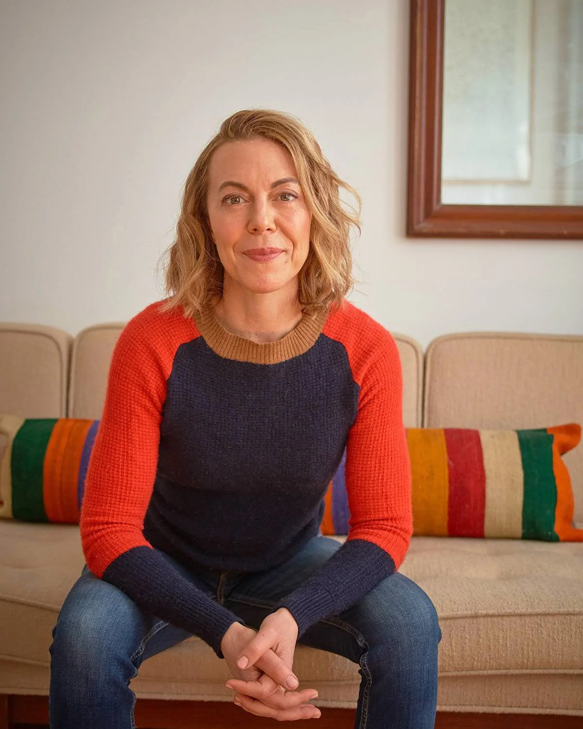 Person sitting on a beige couch with a colorful striped pillow, wearing a sweater with red and navy sleeves, and jeans. A wooden-framed mirror is visible in the background.