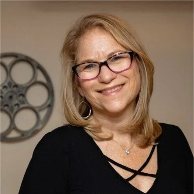 Smiling woman with glasses and blonde hair, wearing a black top, standing in front of a wall with a film reel decoration.