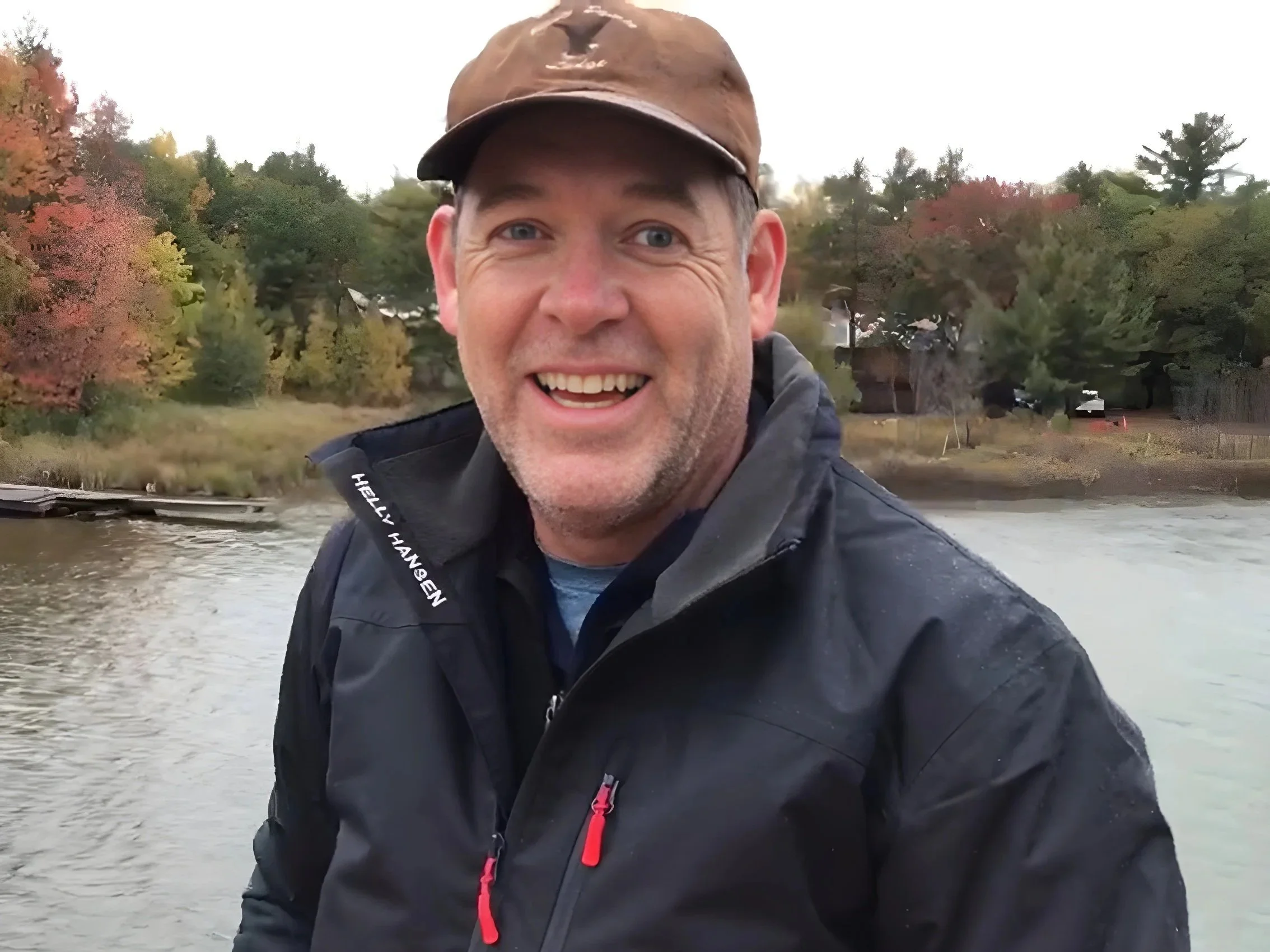 Man smiling outdoors near a body of water with trees displaying fall foliage in the background.