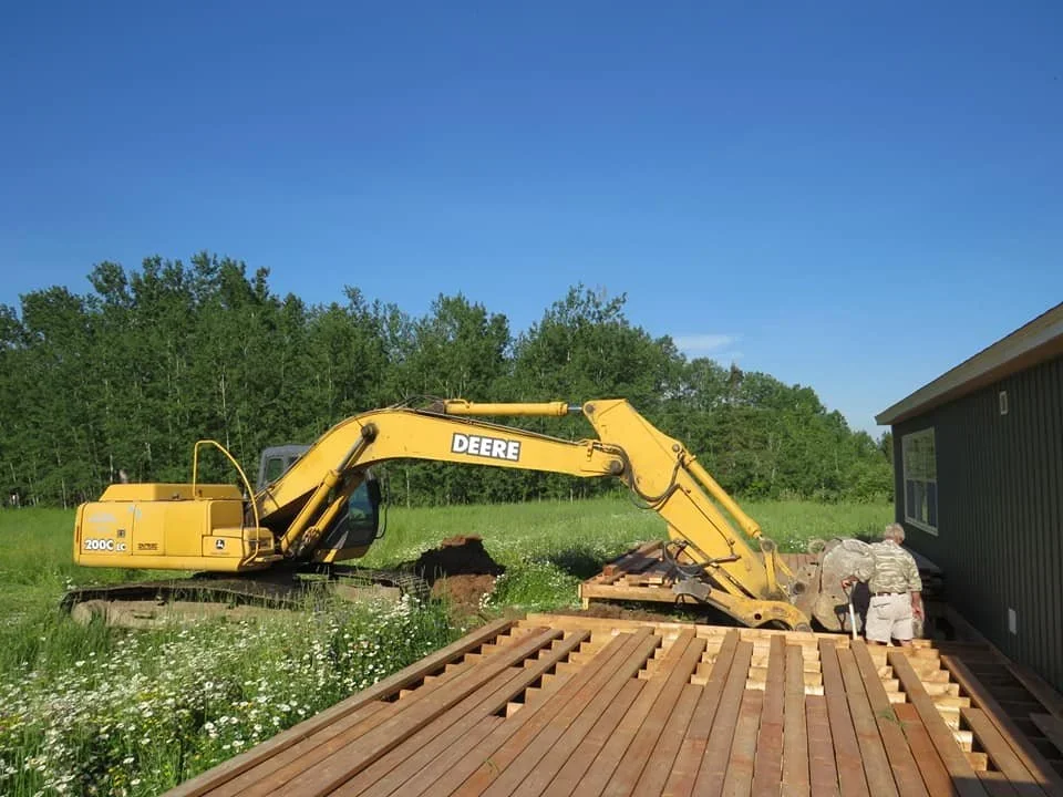 A yellow Deere excavator digging near a house on a grassy field, with wood planks on the ground and a man working near the house.