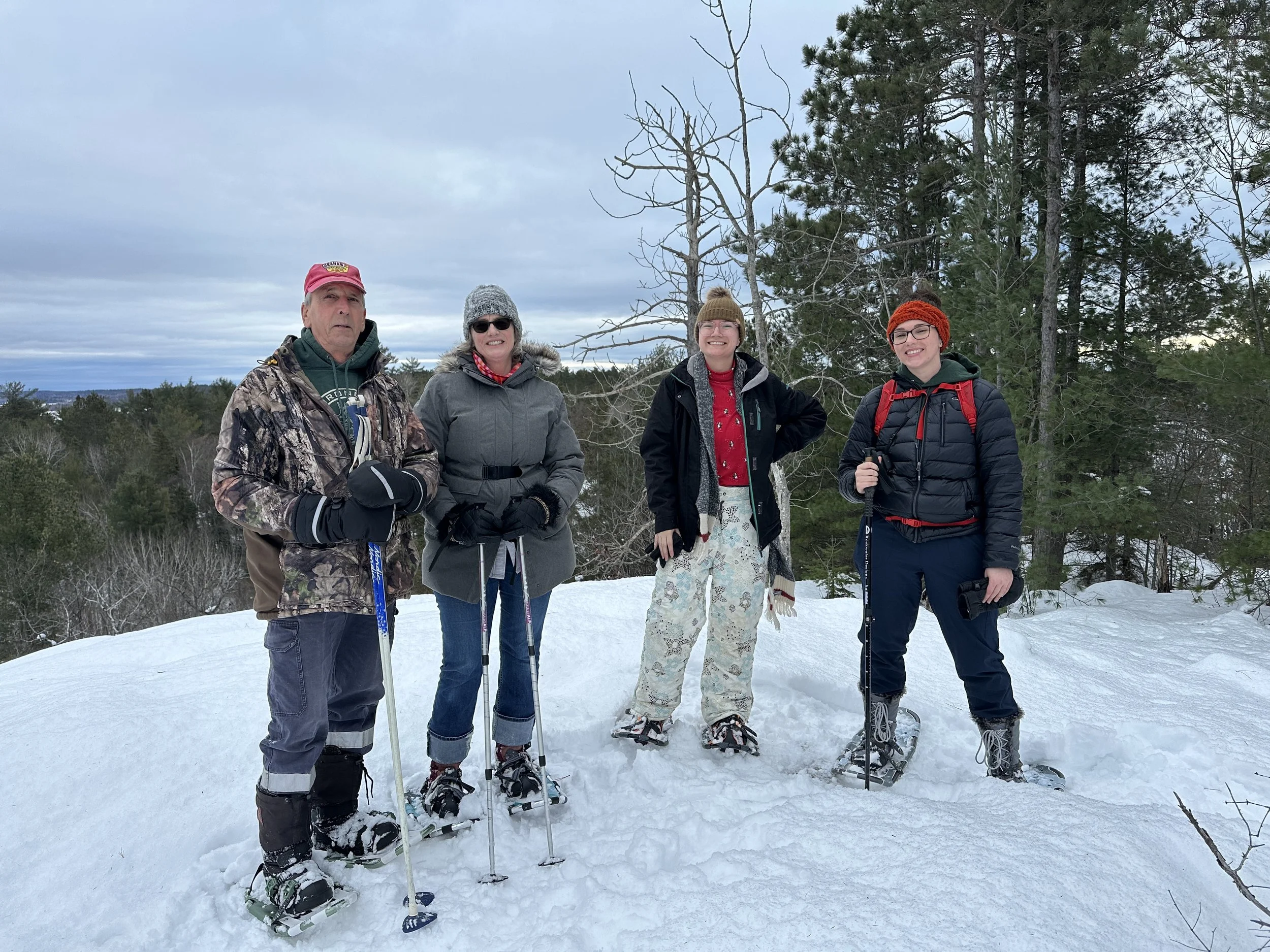 Four people standing in snow with snowshoes, dressed warmly, on a winter landscape with trees and cloudy sky in the background.