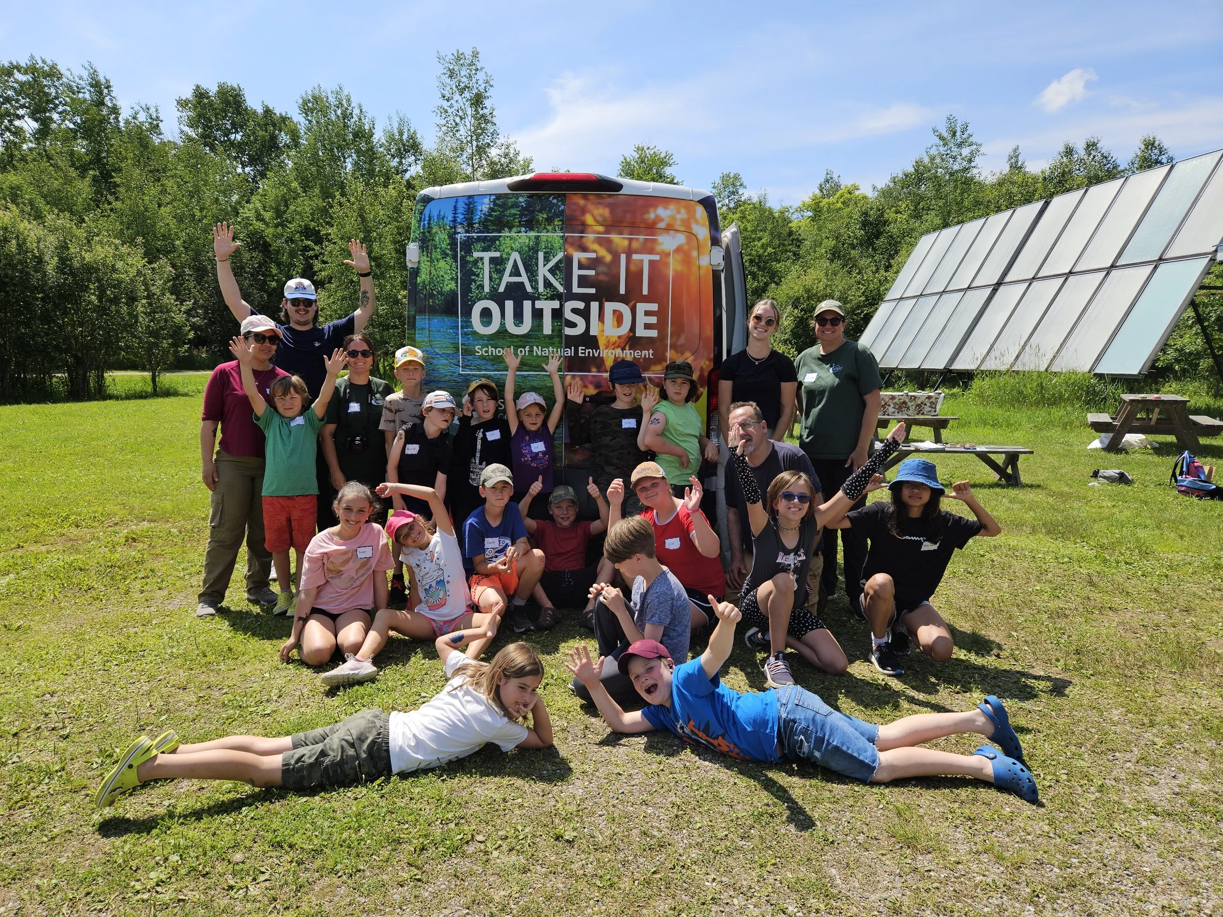Group of children and adults outdoor, posing for a photo in front of a bus with 'Take It Outside' slogan, in a green park with solar panels on the right and trees in the background.