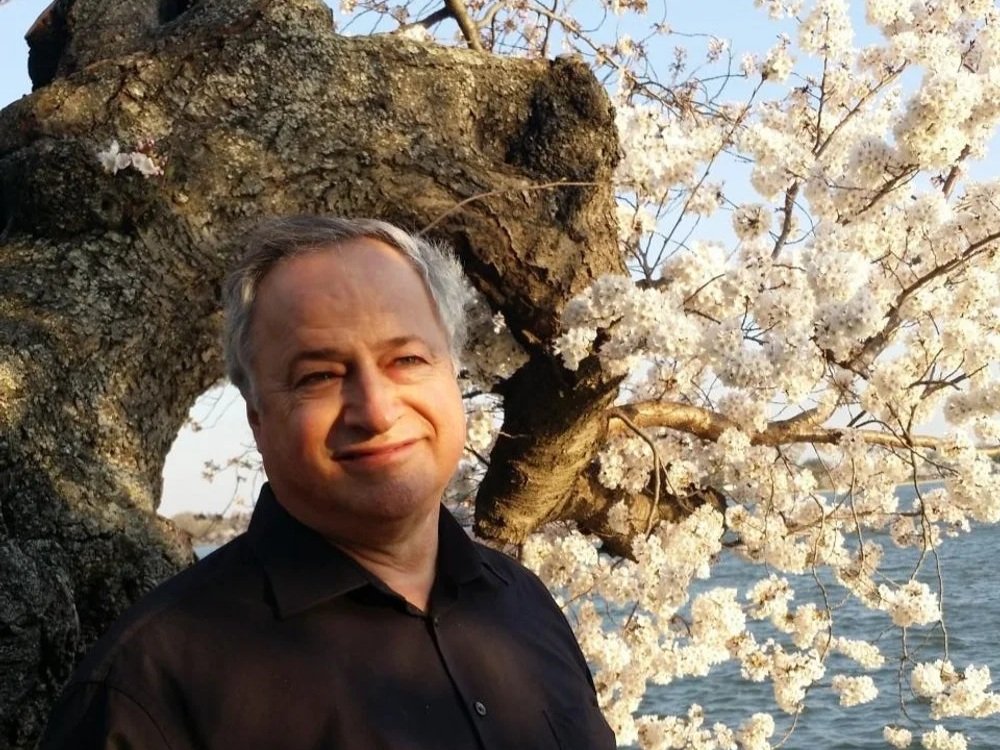 A man stands near a cherry blossom tree with white flowers, next to a body of water, during daylight.