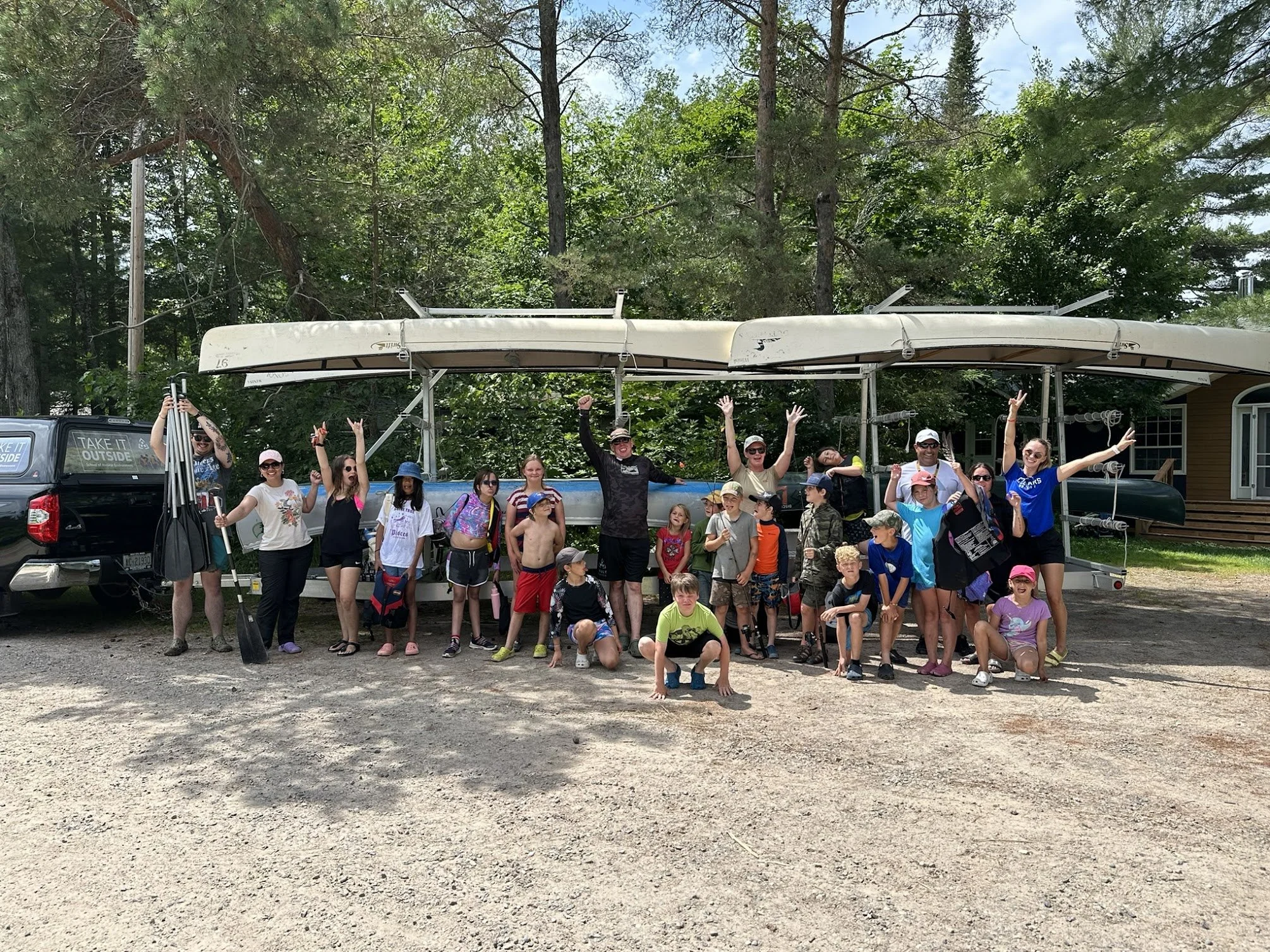 A group of children and adults outdoors, celebrating in front of a boat trailer with canoes and kayaks, surrounded by trees.