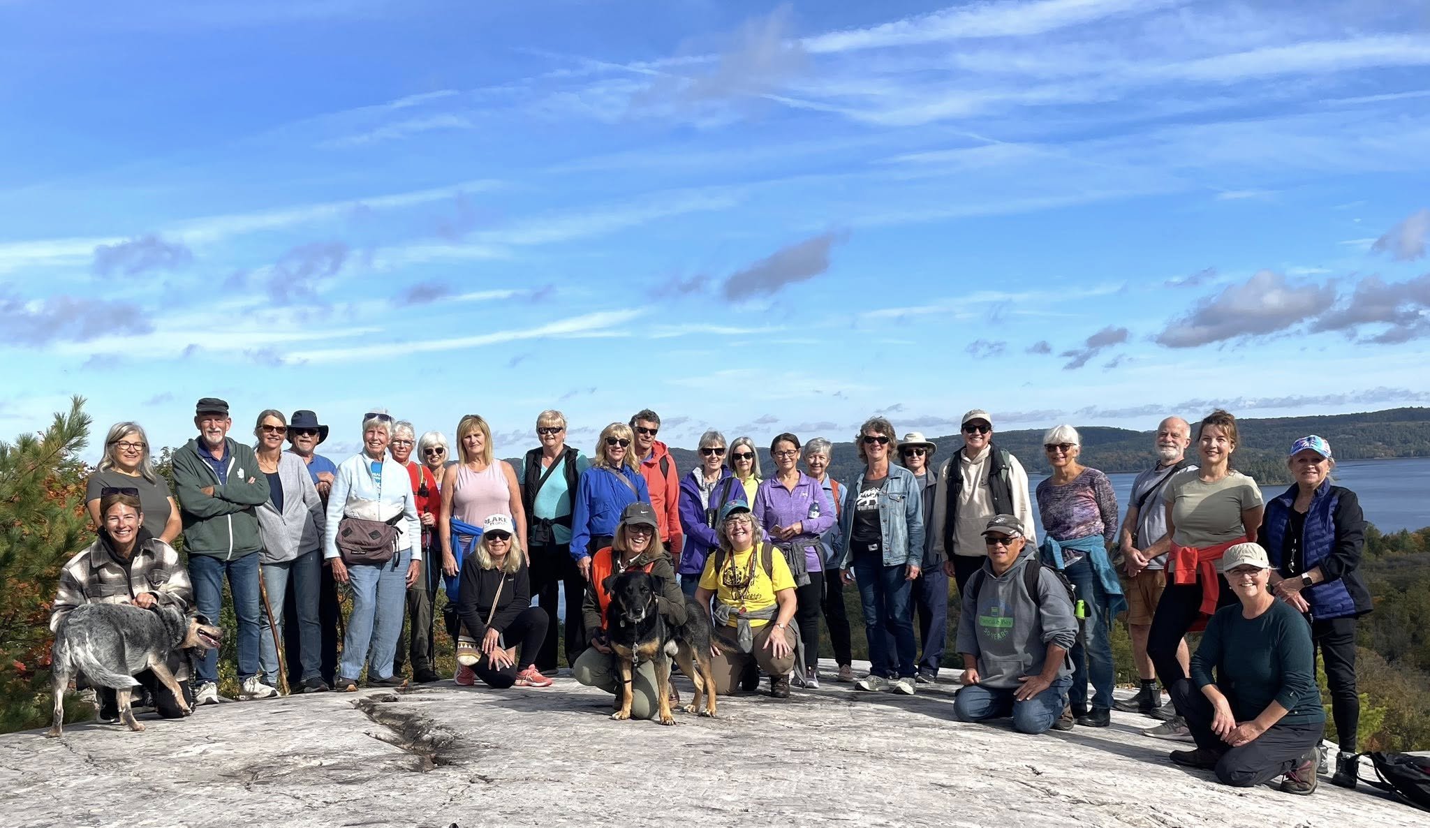Group of people outdoors on a rocky surface with water and hills in the background, some wearing sunglasses and hats, accompanied by dogs, during daytime.