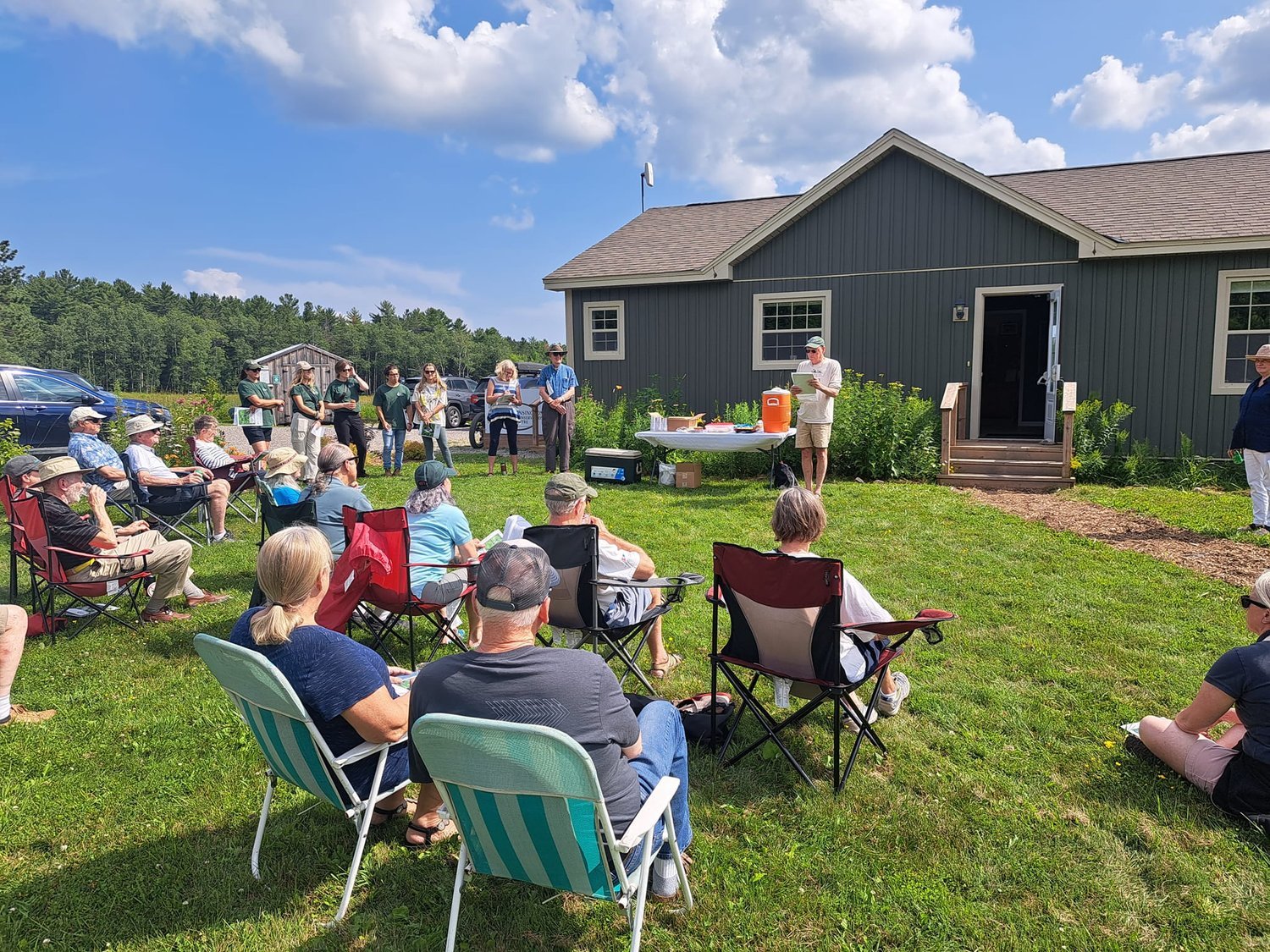 Outdoor gathering at a community event on grass, with people seated in lawn chairs and standing near a building with an open door, under a partly cloudy sky.