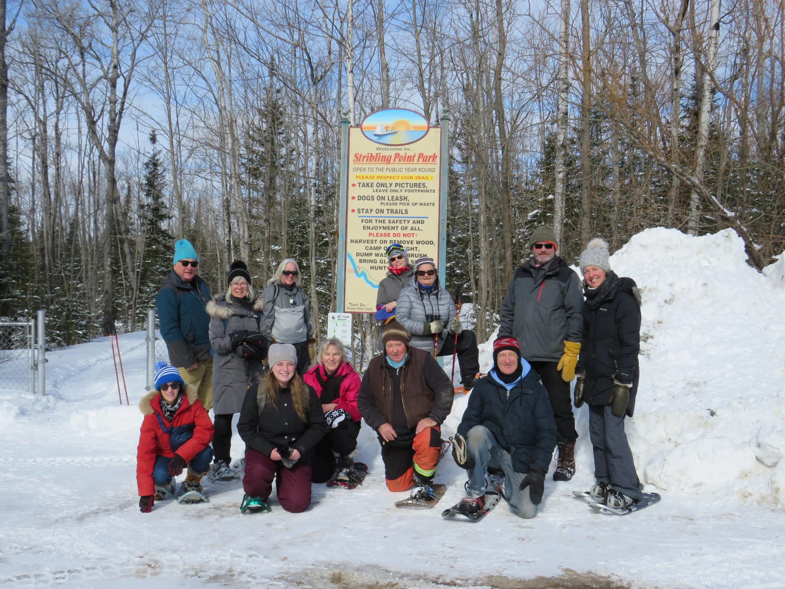 Group of people in winter clothing posing on snow in front of a sign at Stribling Point Park, with a snowbank on the right and leafless trees in the background.