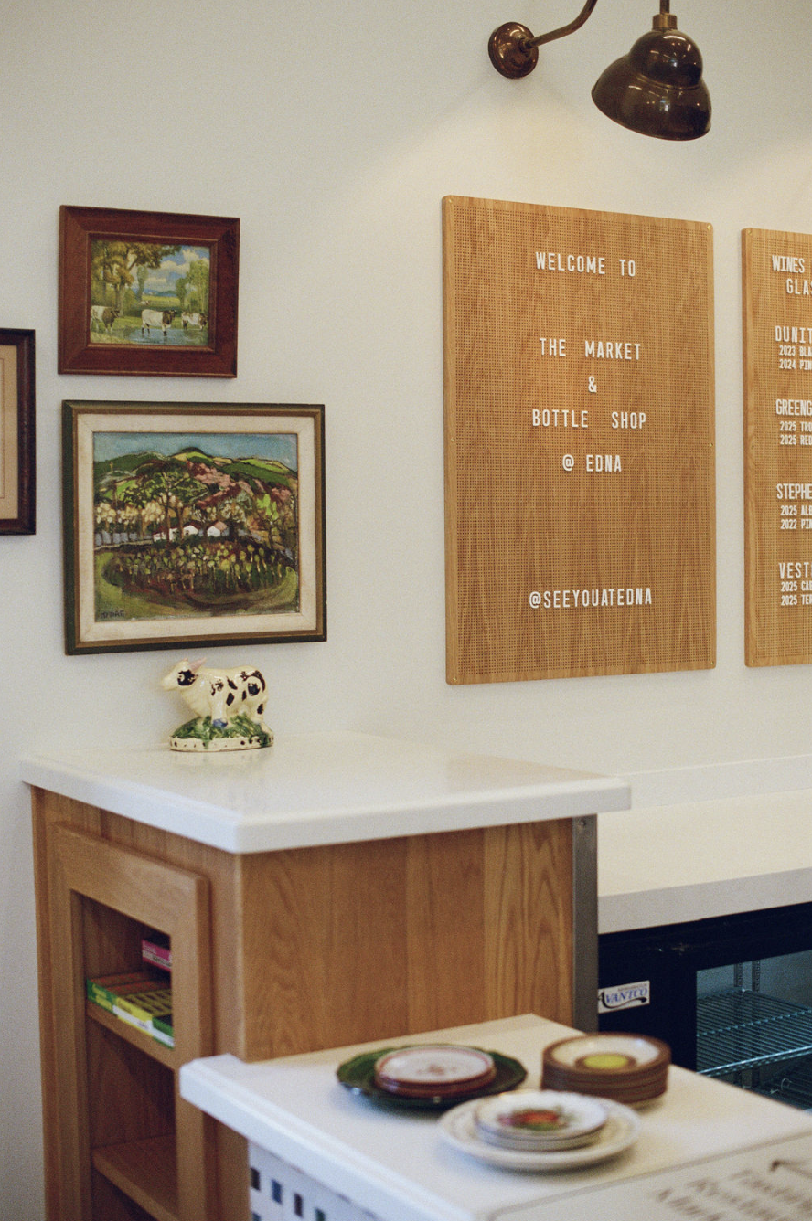 Interior of a shop with framed landscape paintings on a white wall, wooden signs with welcome messages, a ceramic cow figurine, and a table with colorful plates of food.