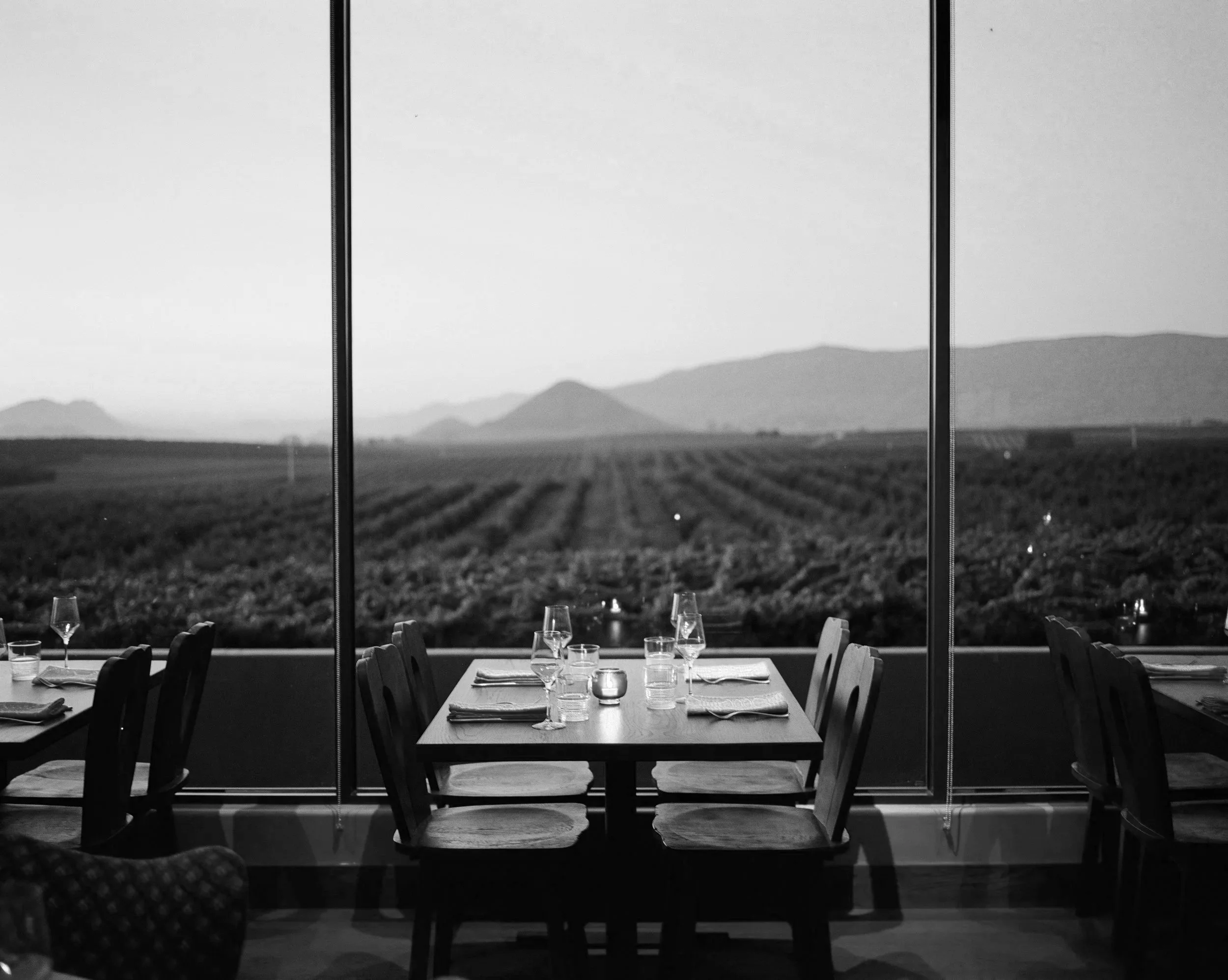 Black and white photo of a restaurant dining area with a large window showing a scenic mountain landscape in the background; tables set with glasses, napkins, and candles.