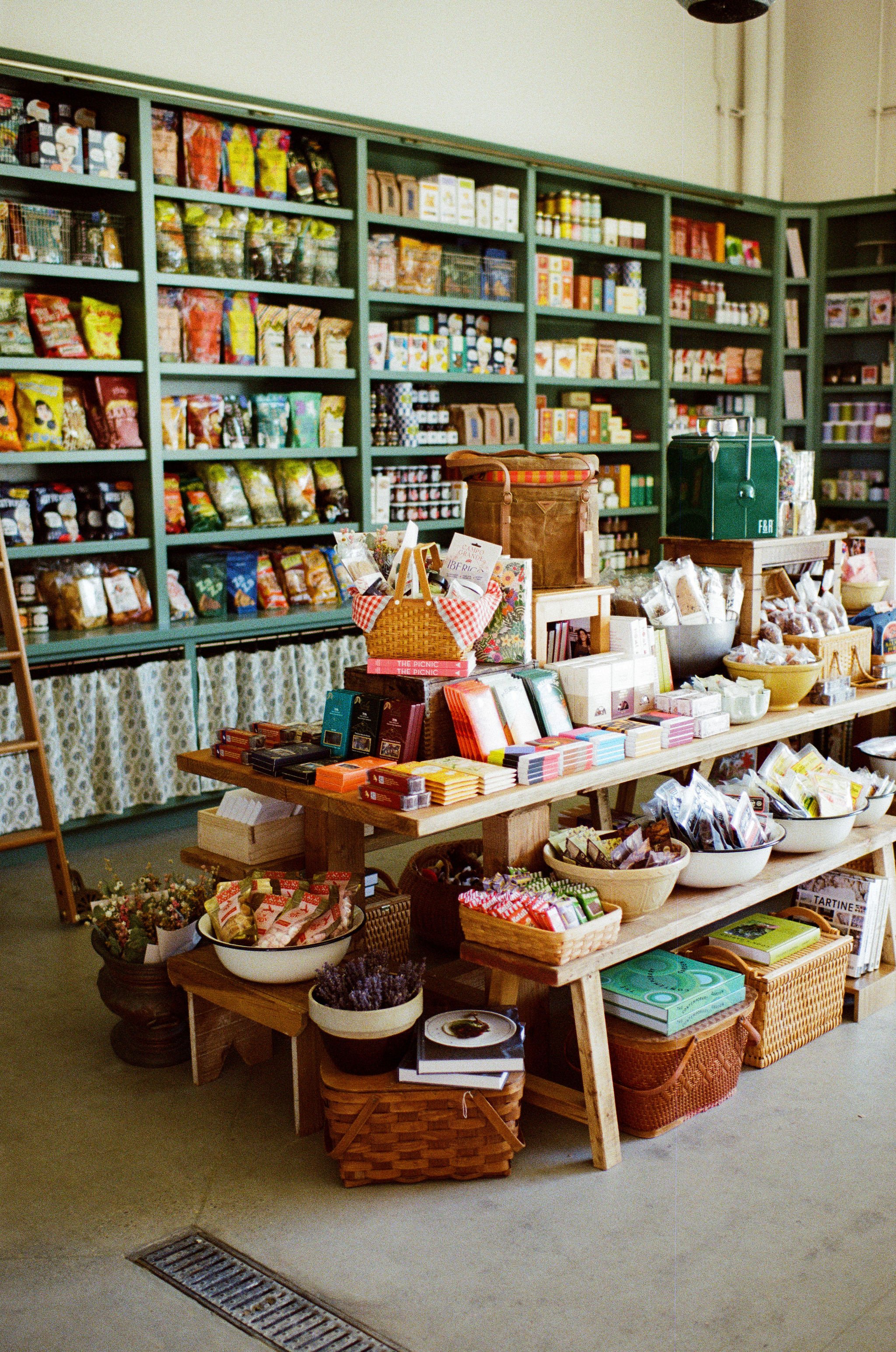 Interior of a shop with shelves filled with various packaged food items, small tables with baskets and boxes of assorted products, including books, small decorative plants, and baskets of candies or snacks.