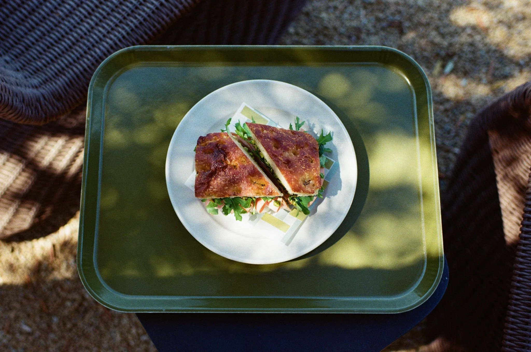 A sandwich with meat, lettuce, and tomato on a white plate, placed on a green tray outdoors.