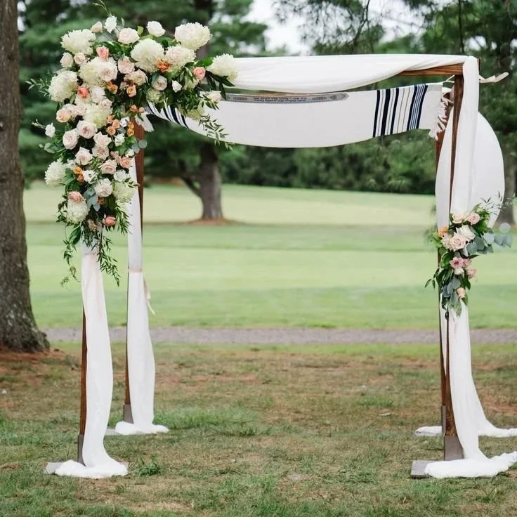 Outdoor wedding arch decorated with white and pink flowers and draped white fabric, set in a grassy area with trees in the background.