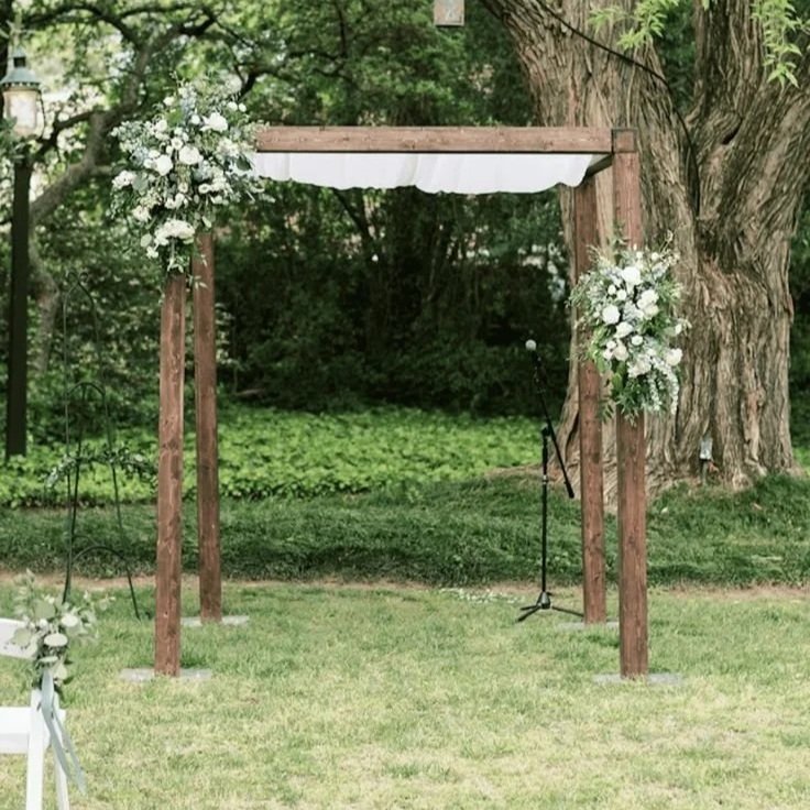 Outdoor wedding ceremony setup with a wooden arch decorated with white flowers, planted in a grassy area with trees in the background.