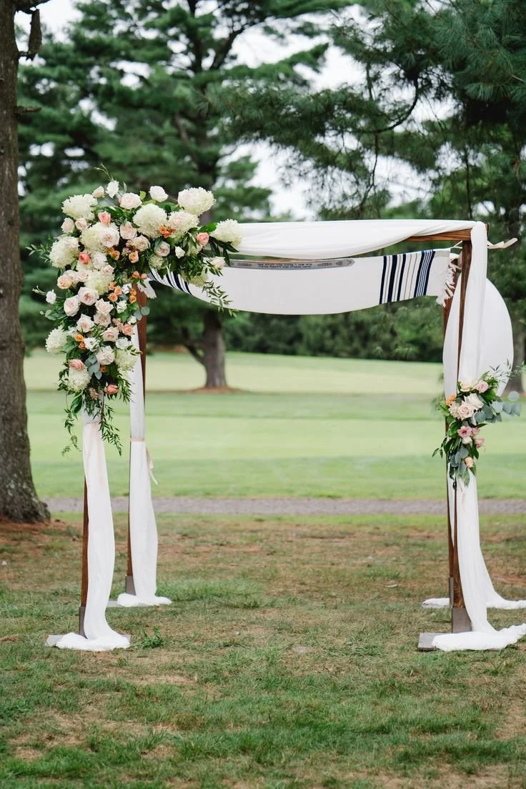 Outdoor wedding arch decorated with white and pink flowers and draped white fabric, set in a grassy area with trees in the background.