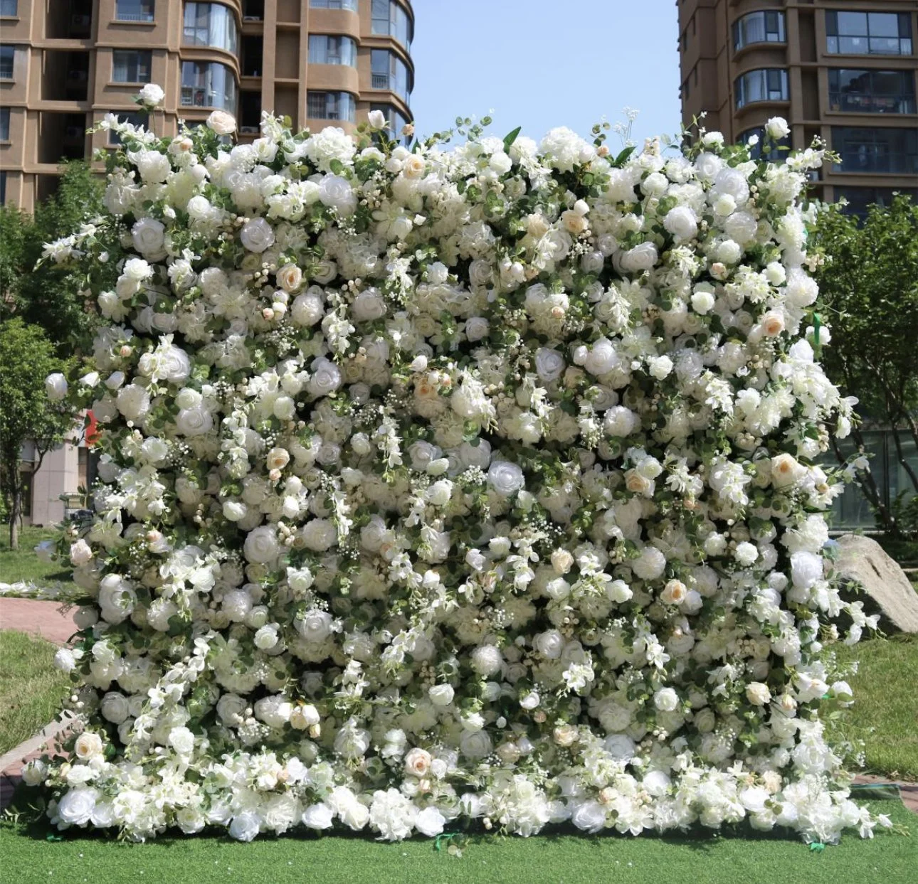 A large cluster of white flowers, including roses, is arranged as a decorative display in an outdoor setting with grass and trees, and modern buildings in the background.
