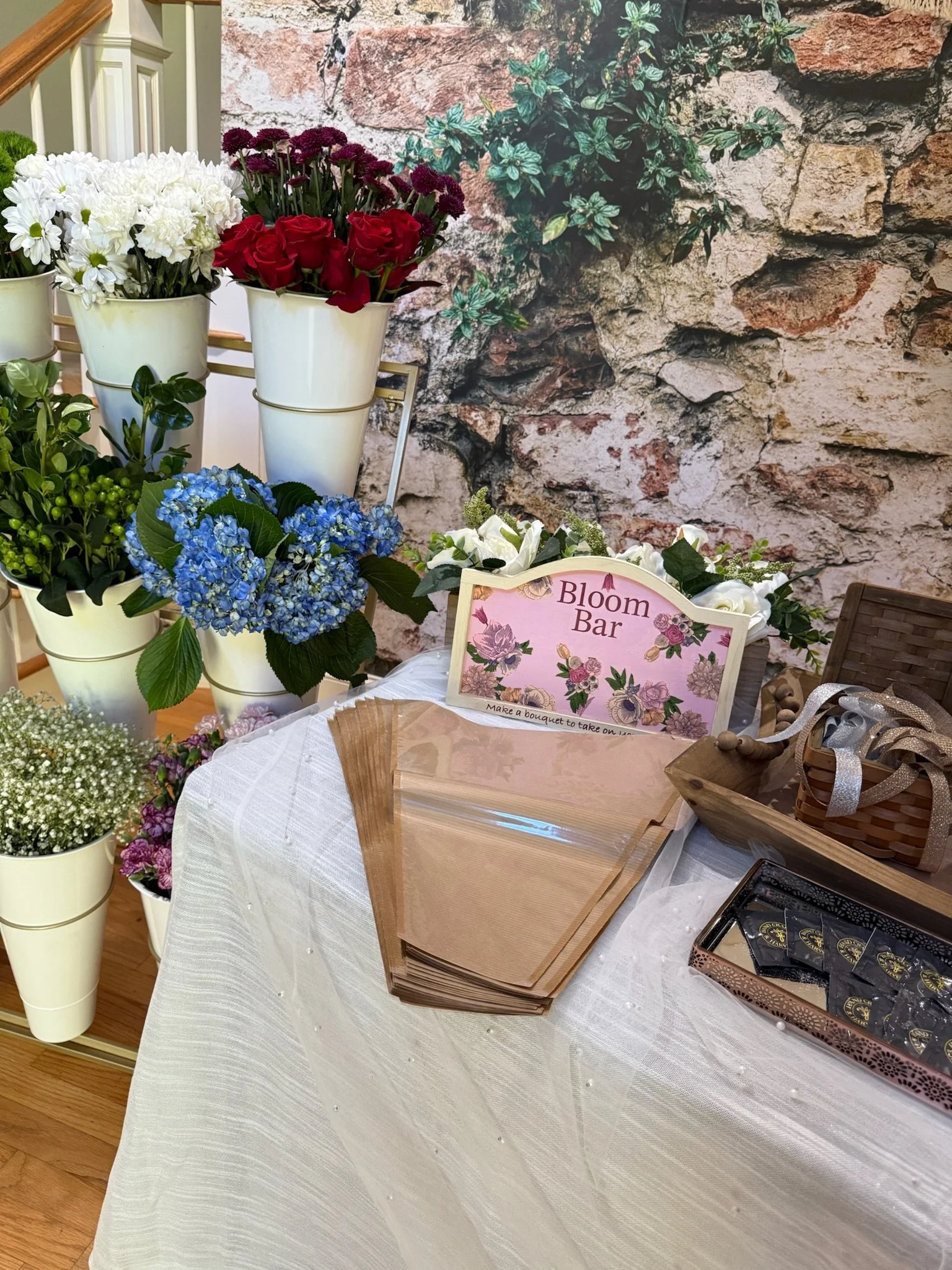Display of various flowers in white pots, including white daisies, red roses, blue hydrangeas, and baby's breath, with a sign that reads "Bloom Bar" on a table covered with a white tablecloth, against a brick wall background.