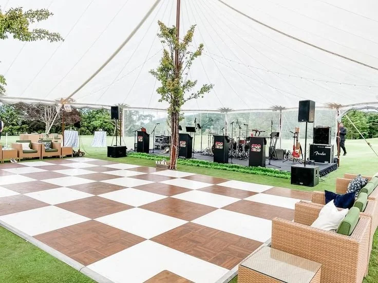 Outdoor tented event space with a checkered dance floor, musical instruments on a stage, and seating areas with couches and pillows.